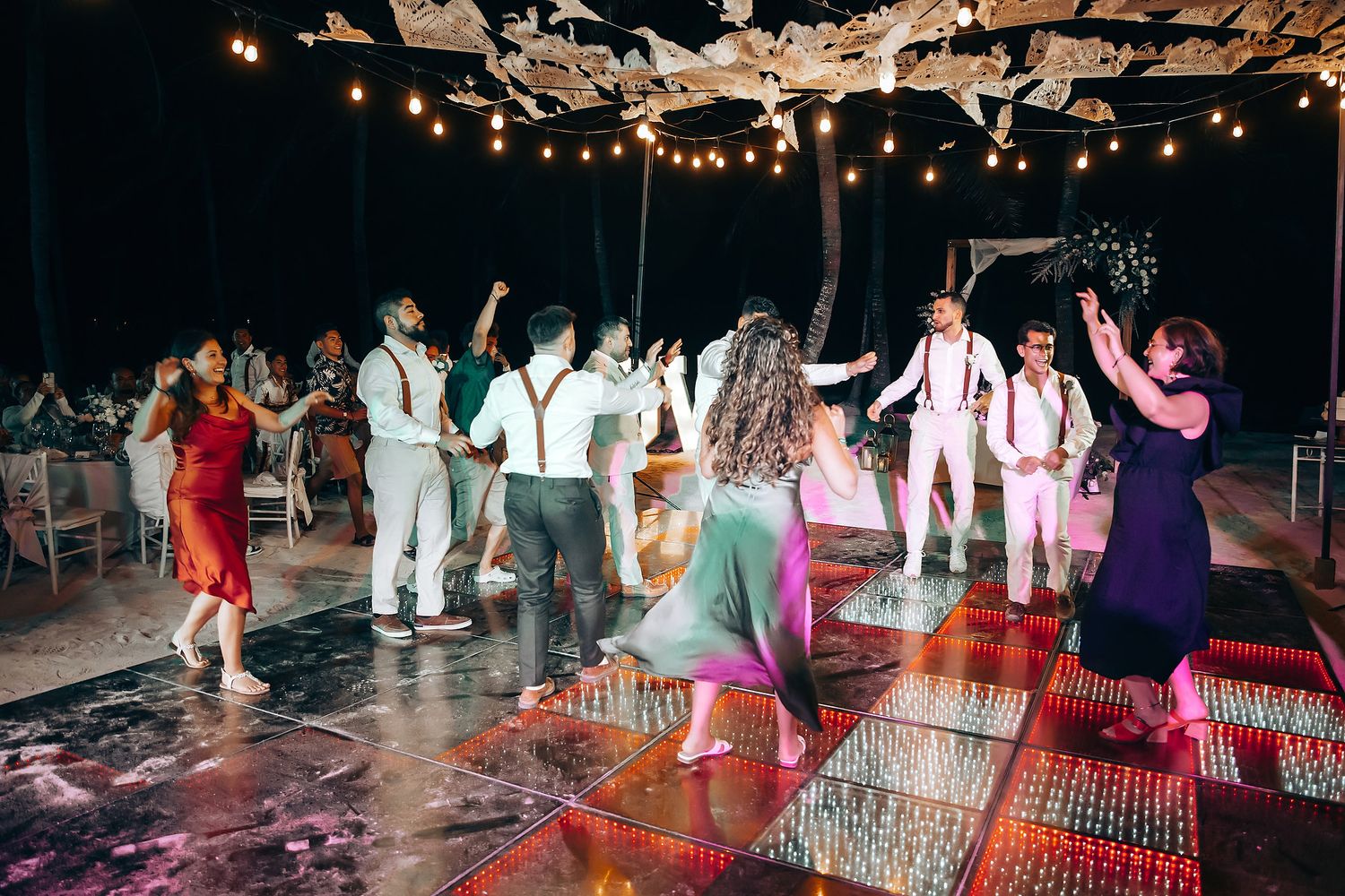 Wedding reception guests dancing under twinkling lights on a tropical beach at night.