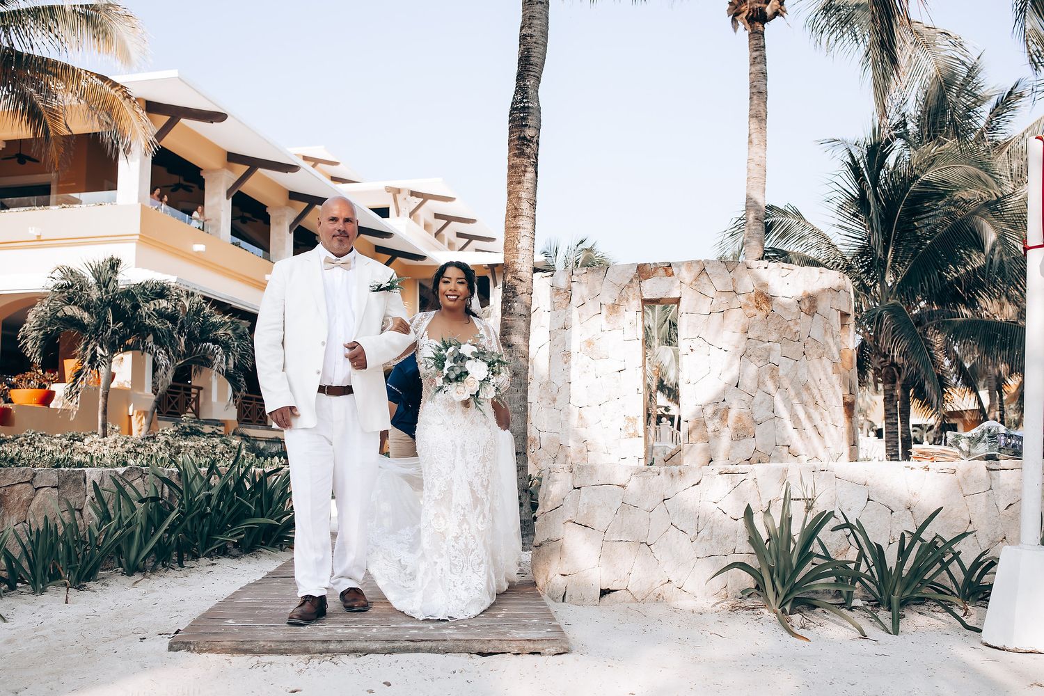 Elegant wedding photos captured along a palm tree-lined beach path with white and beige architectural elements.
