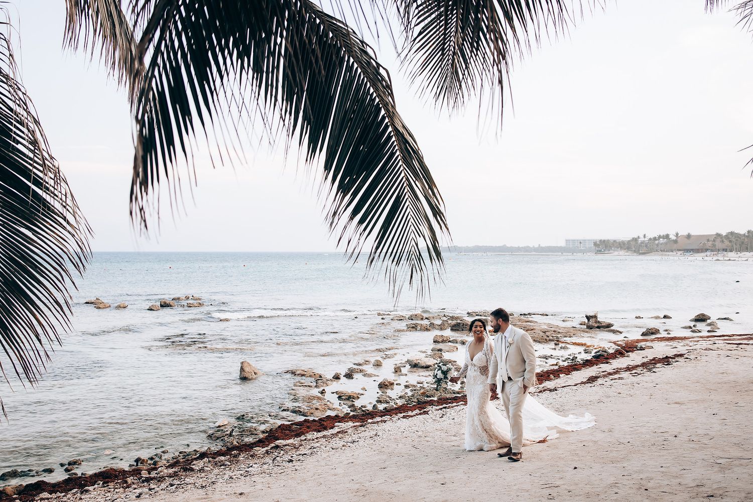 Couple walking along a tropical beach with palm trees and ocean views at sunset.