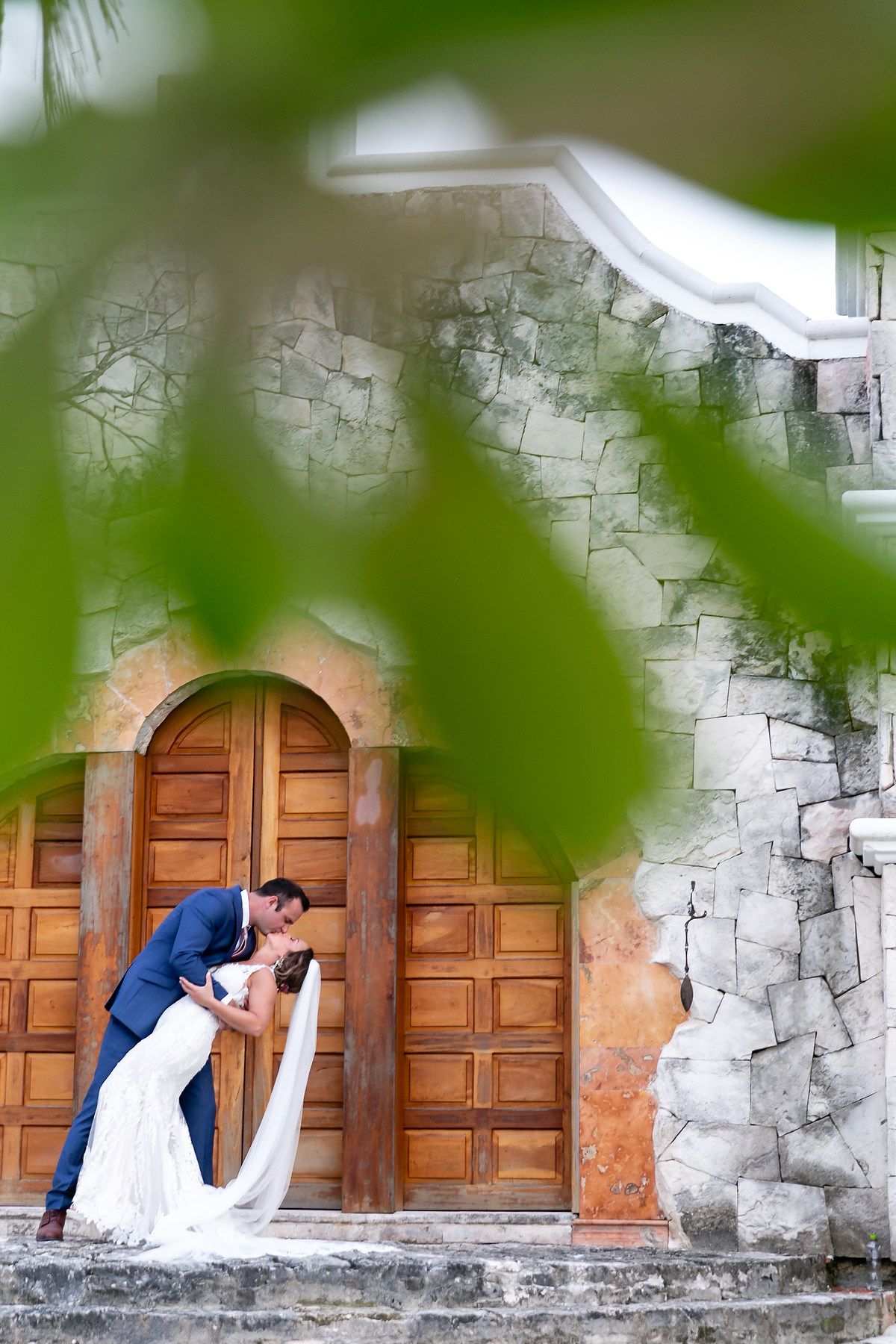 A romantic wedding moment captured through tropical foliage at a historic stone church with wooden doors.