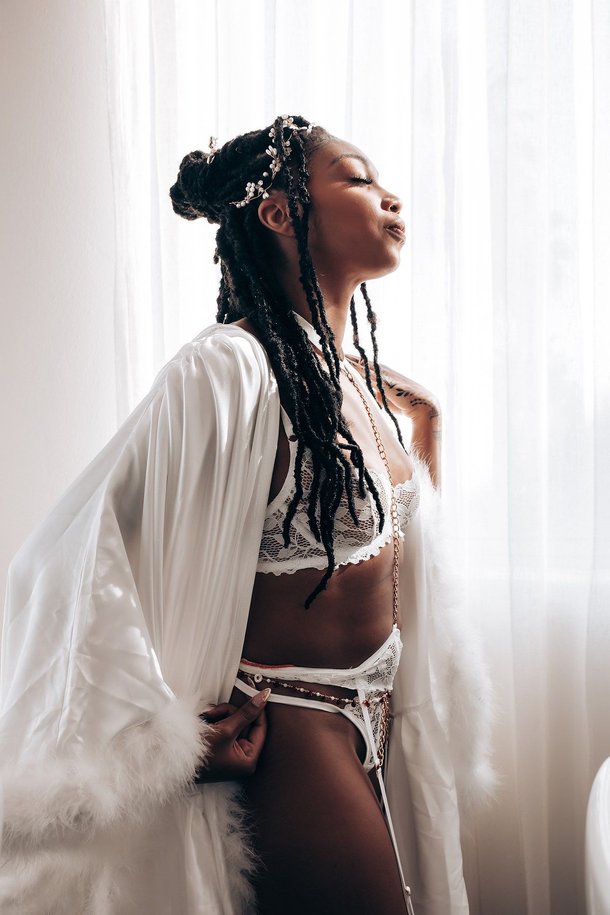 A woman in white lingerie and robe poses elegantly against a bright window with braided hair adorned with beads.