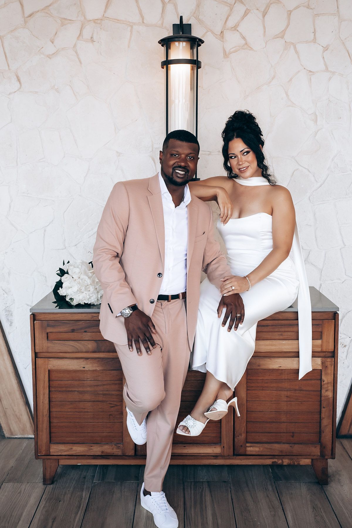 A couple poses playfully on a wooden bench in coordinated formal wear of pink suit and white dress.