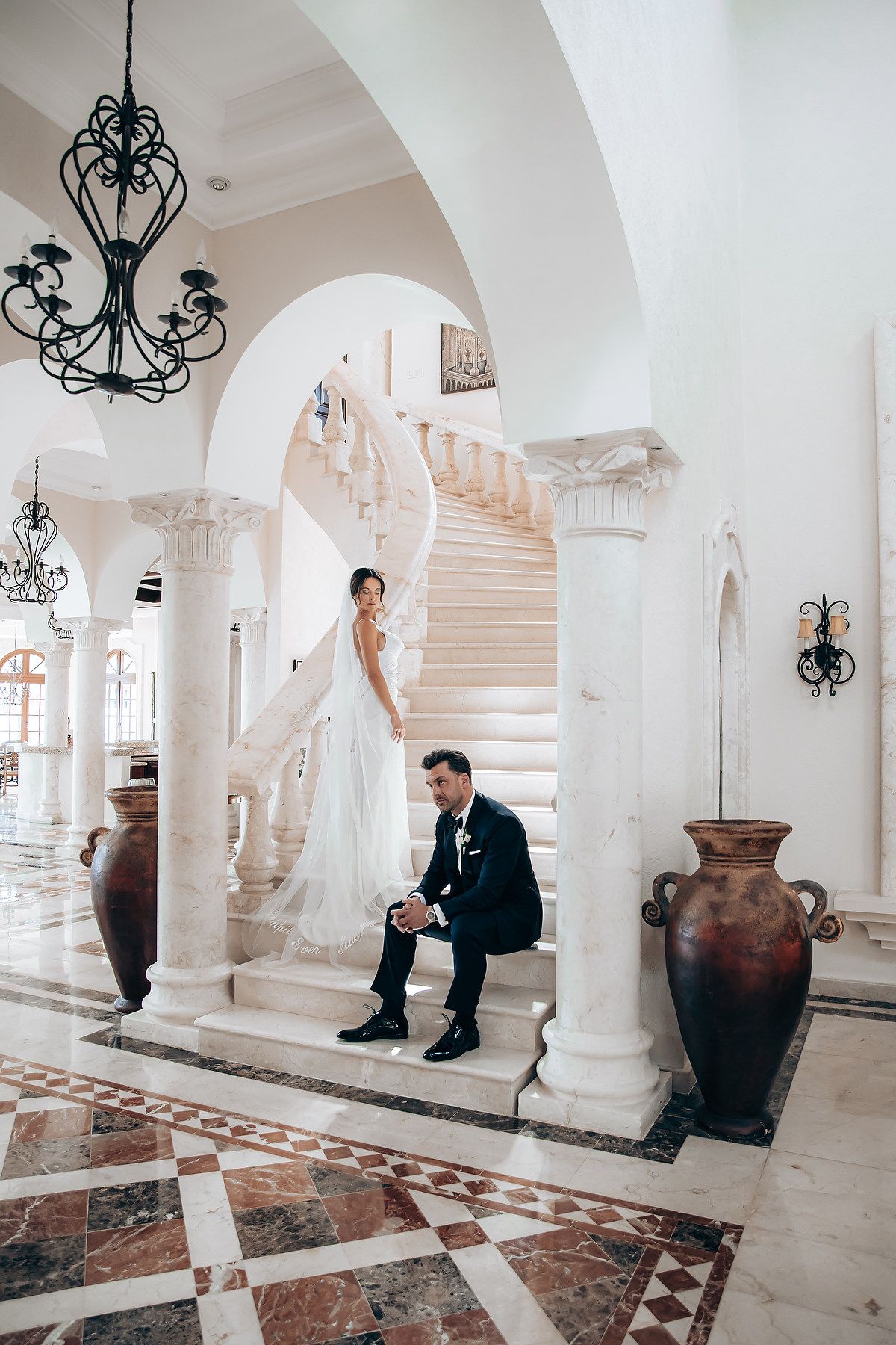 Groom sits thoughtfully on marble steps in an elegant white hallway with wrought iron chandeliers.