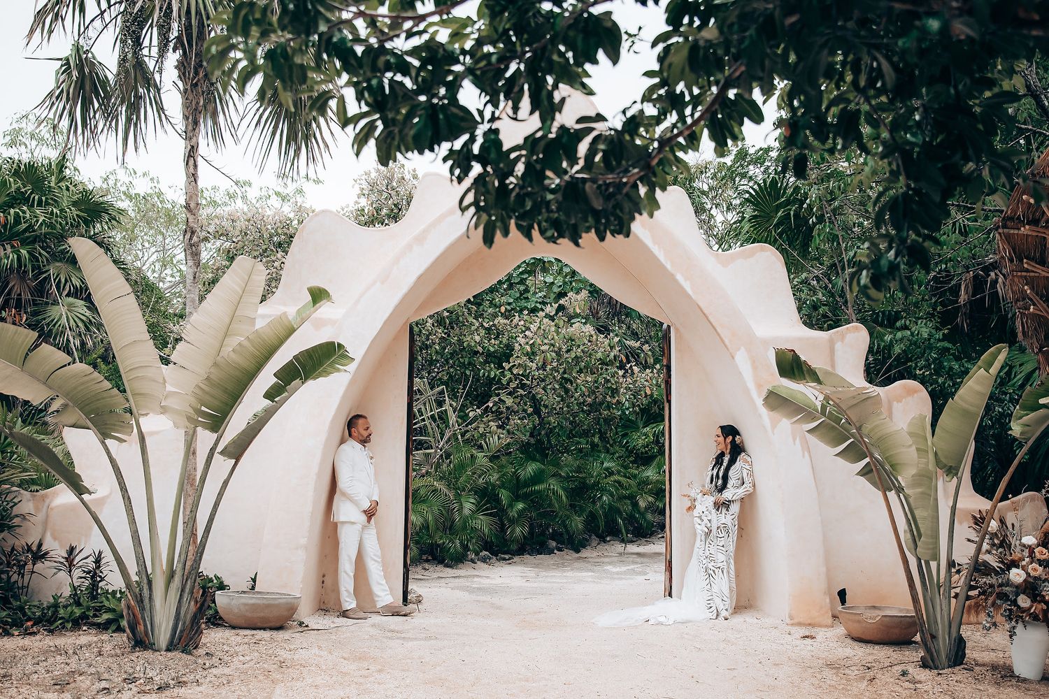 Beachside wedding ceremony under a floral arch with palm trees and ocean view.