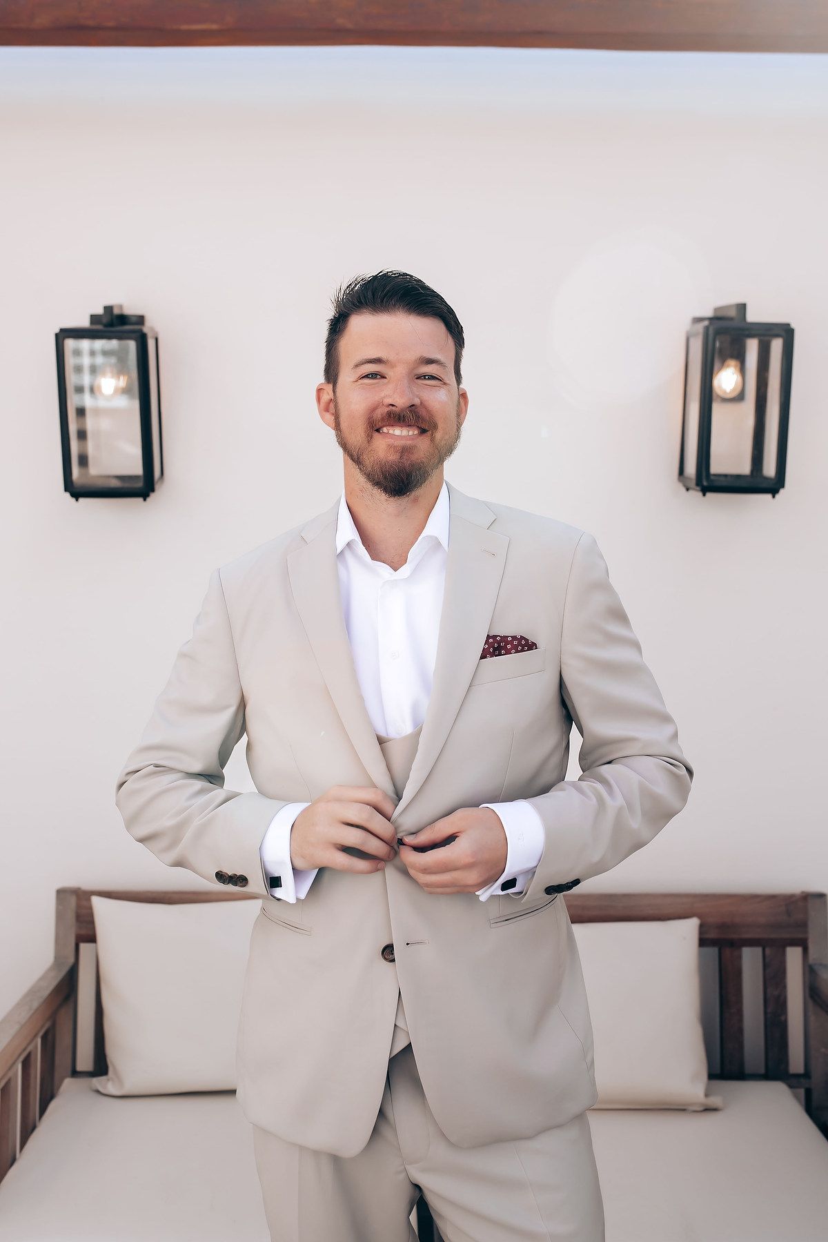 Groom in light beige suit and white shirt poses confidently against white wall with wooden bench.