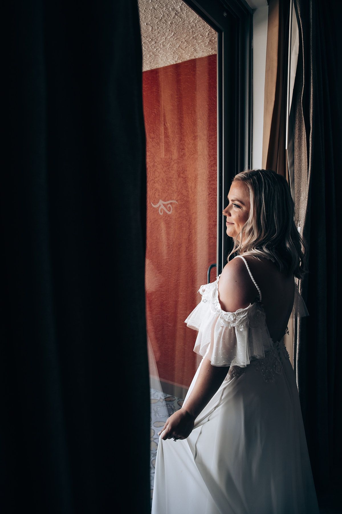 Woman in white dress poses elegantly by window with dramatic lighting and curtains in moody indoor setting.