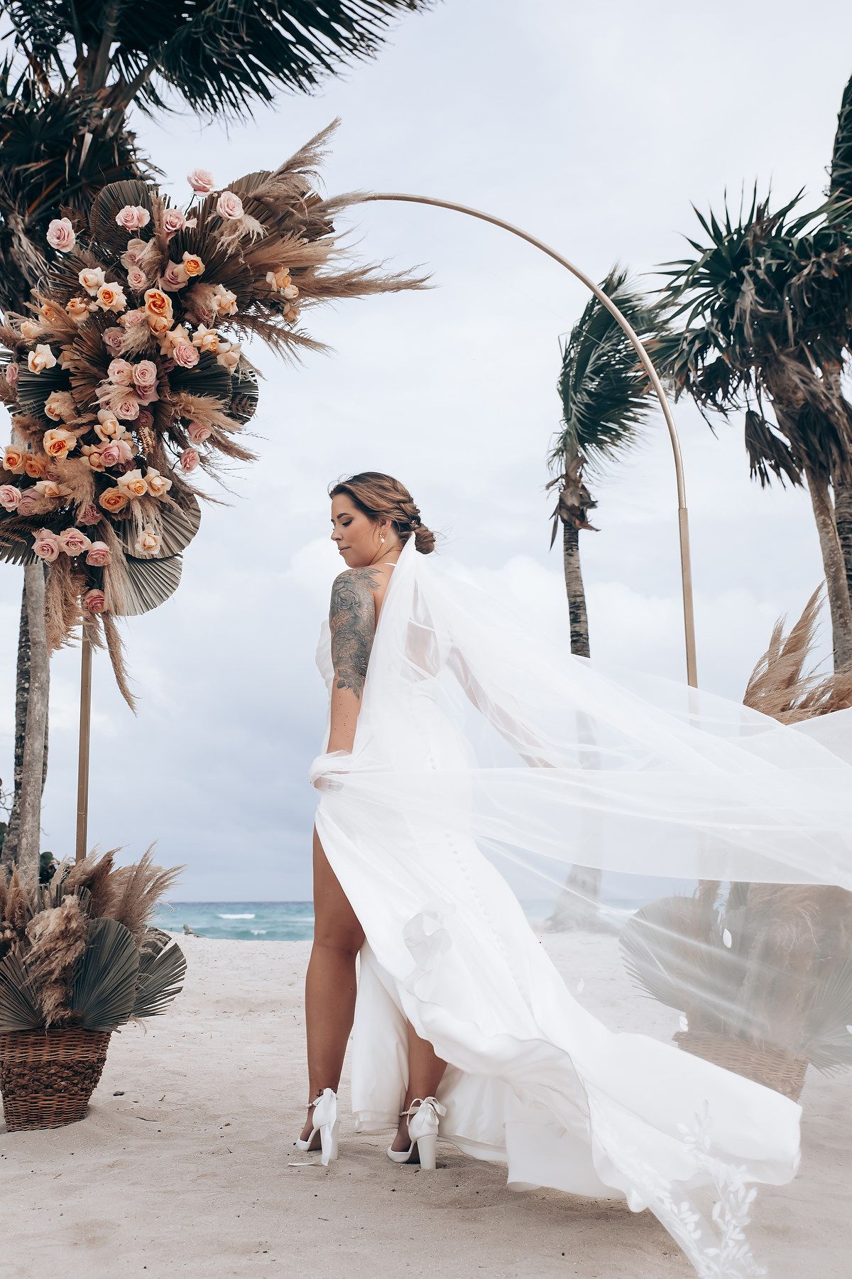 A flowing white wedding dress billows in the ocean breeze along a tropical beach lined with palm trees.