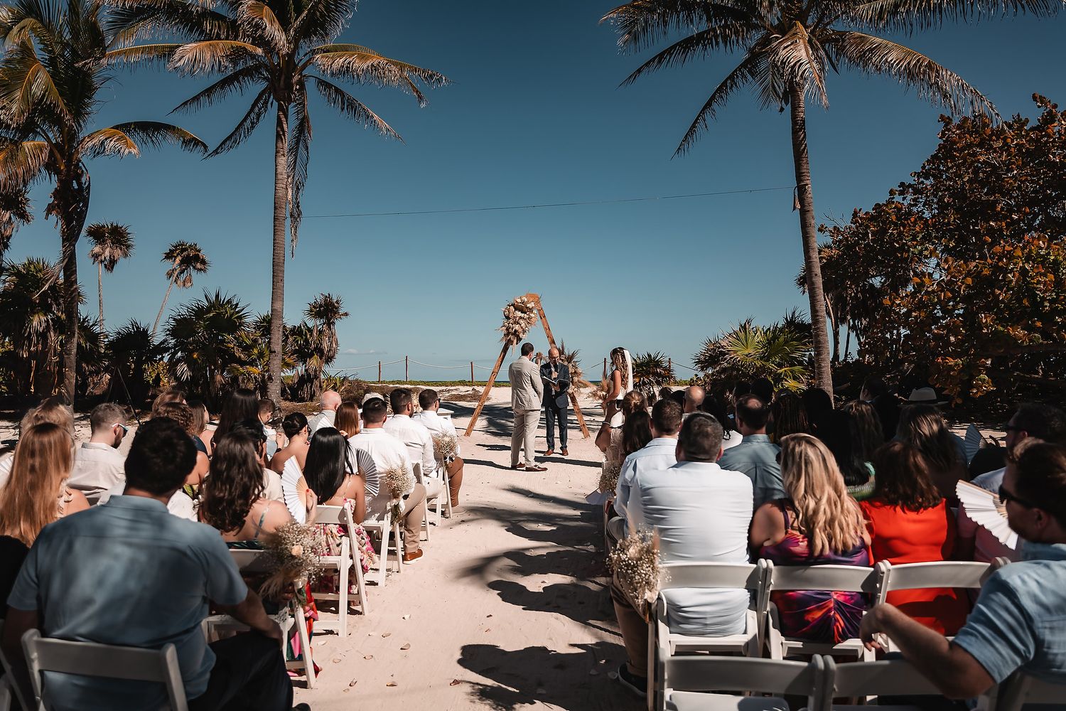 Beach wedding ceremony setup with white chairs and palm trees against a bright blue sky.