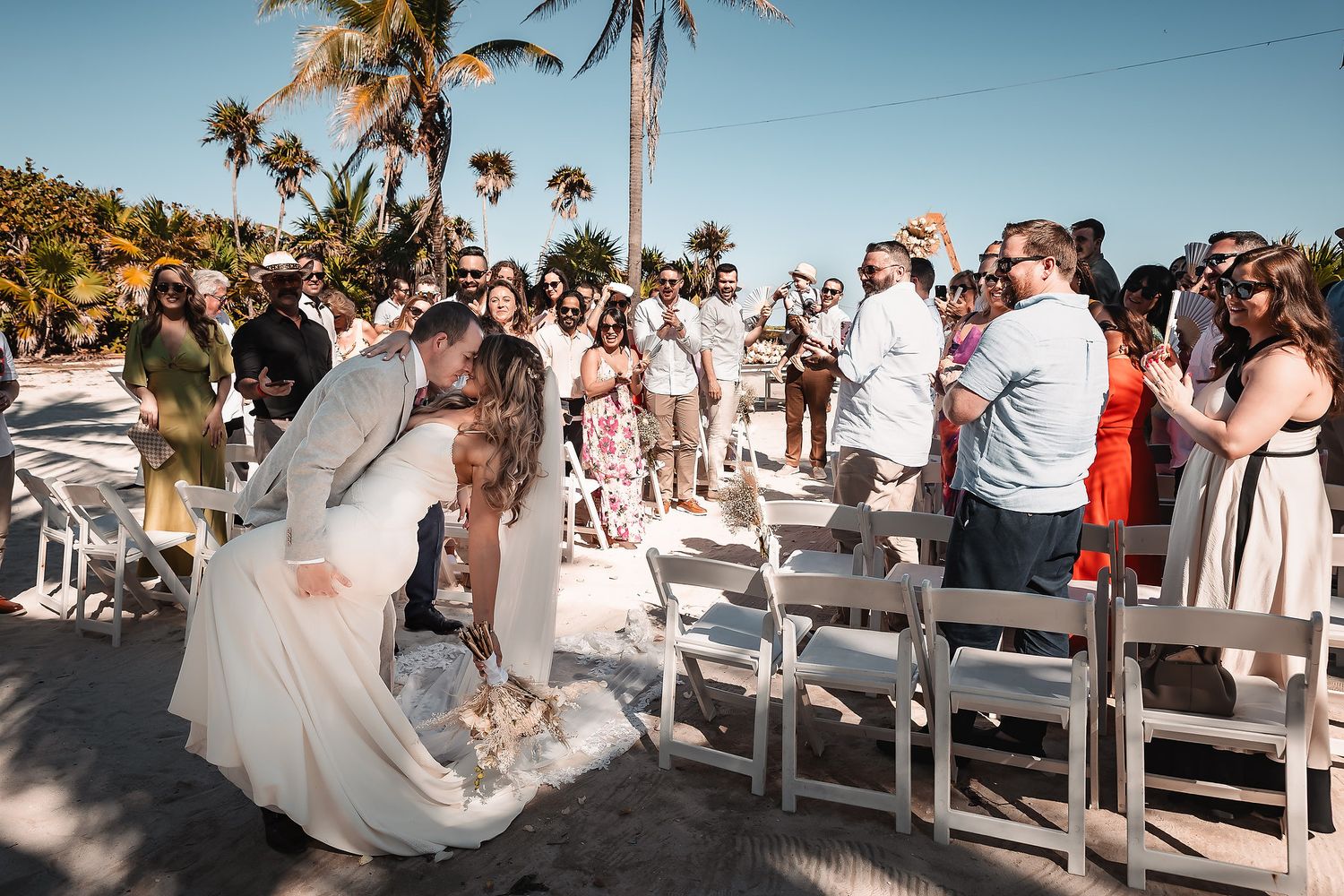 Wide angle shot of wedding guests seated in white chairs along a tropical beach path.