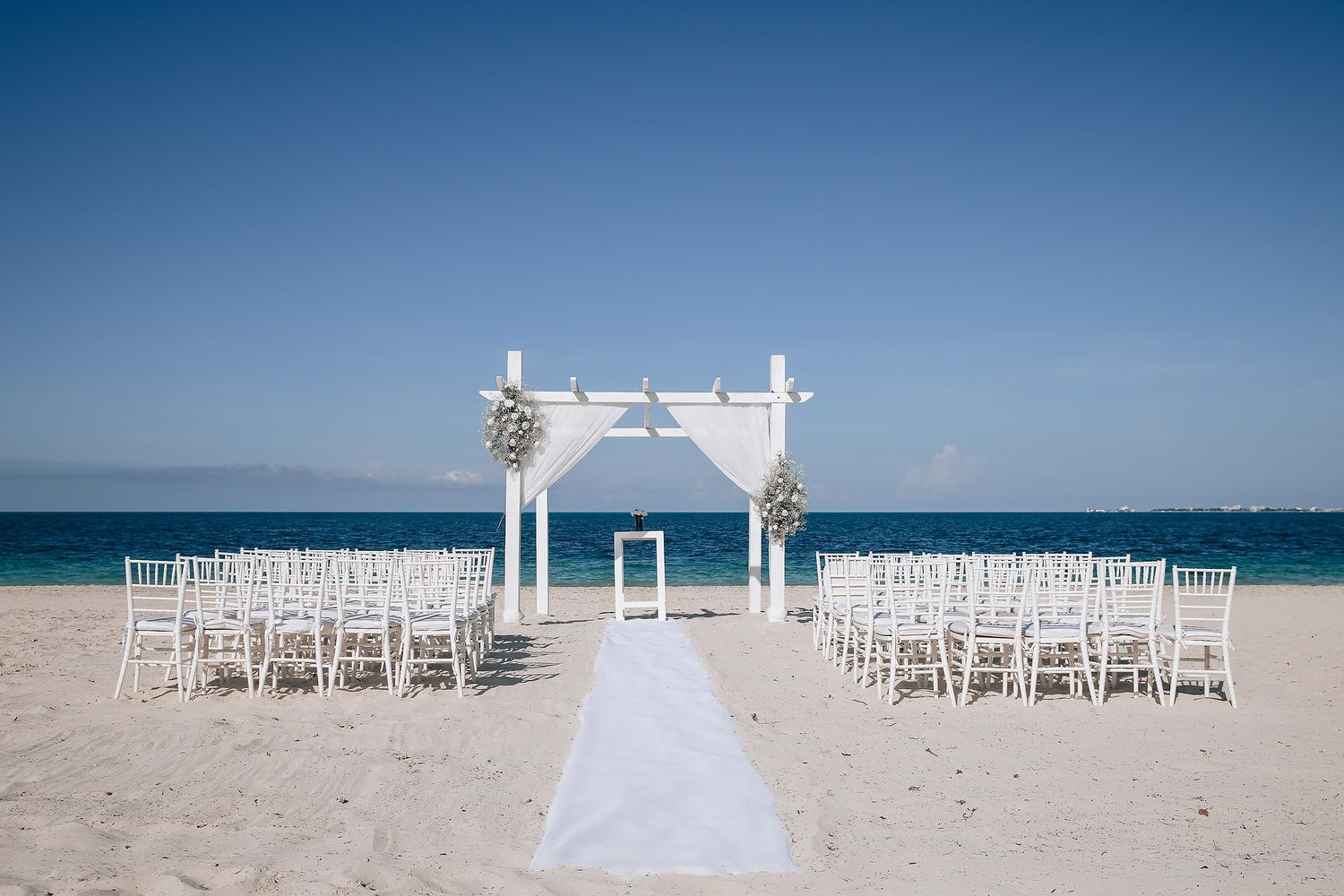 Rows of white chairs arranged symmetrically on a sandy beach with ocean view and wedding arch.