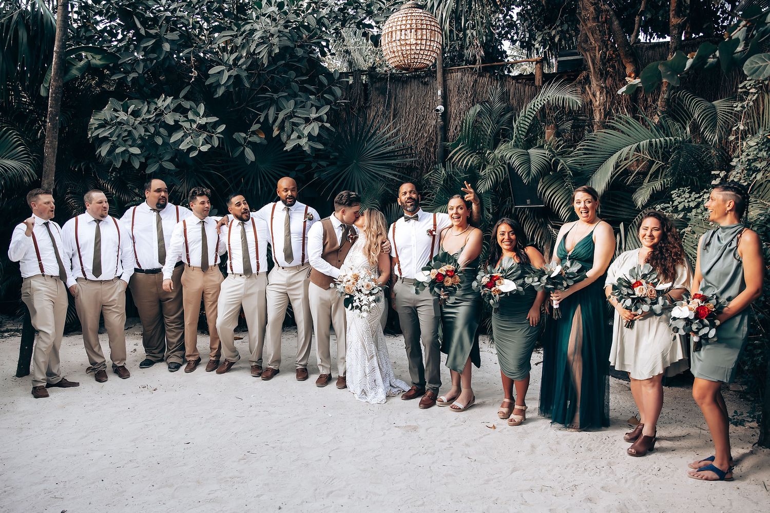 Wedding party in coordinated attire of white shirts, suspenders and teal dresses pose together in an outdoor garden setting.