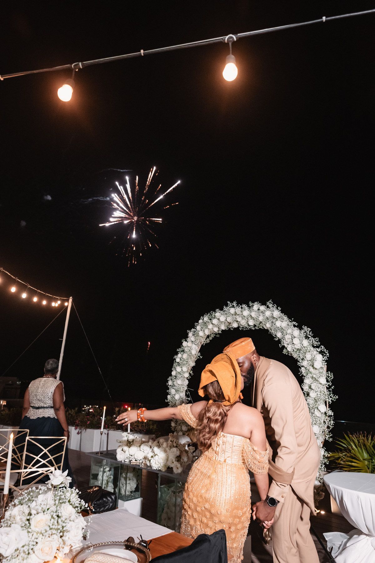 Couple cuts wedding cake under fireworks and string lights at an outdoor nighttime reception celebration.