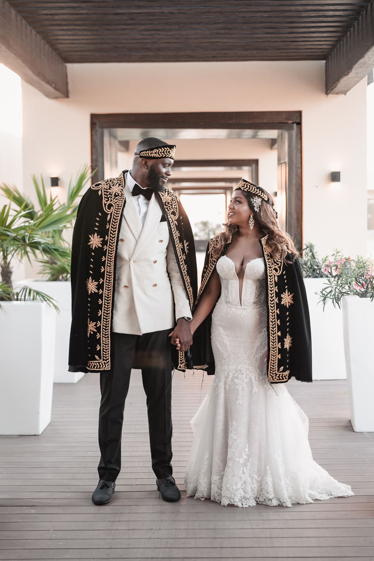 Couple in elegant African-inspired royal wedding attire with ornate black and gold embroidered capes hold hands.