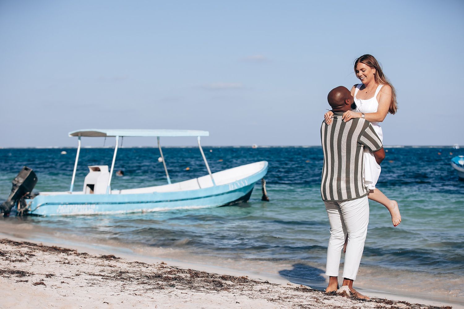 Romantic couple embracing on a tropical beach with a blue boat in the background on a sunny day.