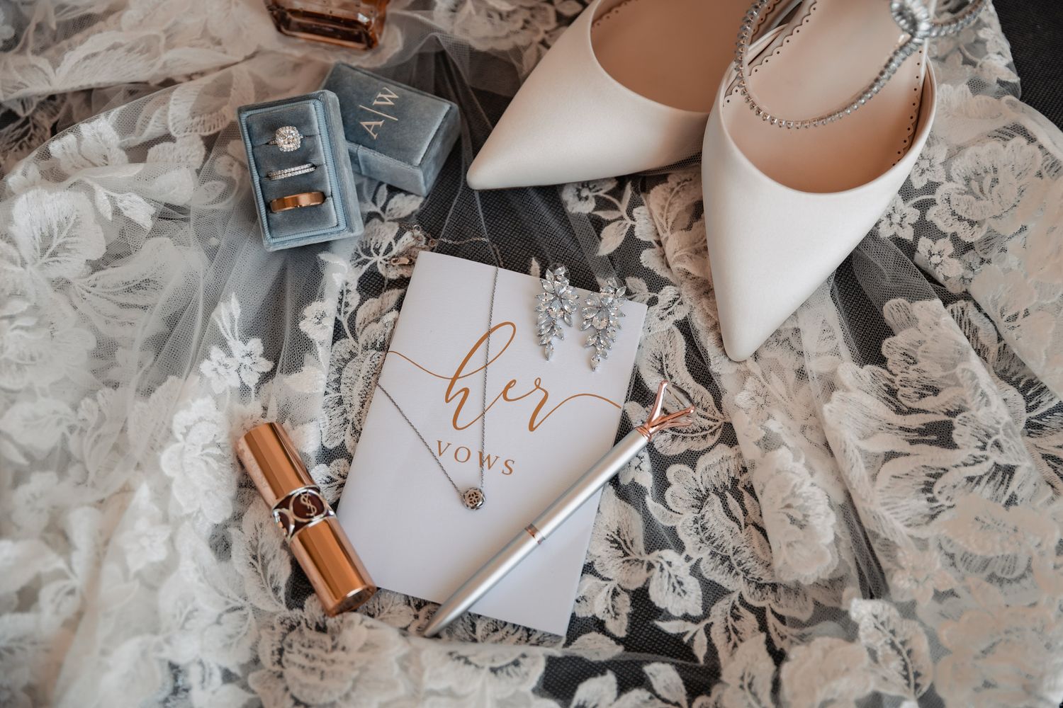 Bridal flat lay with white heels, vow book, ring boxes, lipstick, and floral lace fabric on wedding day.
