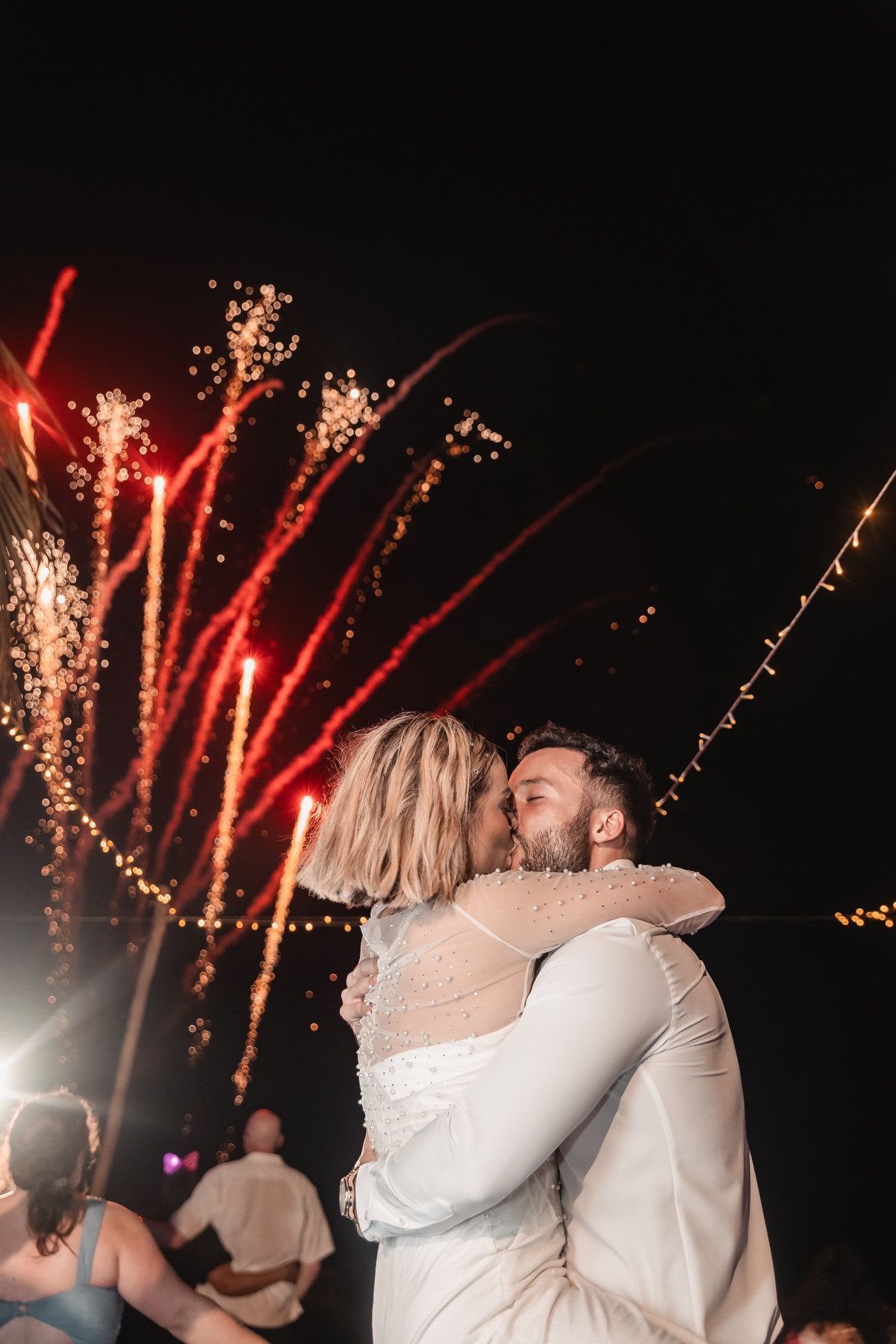 Newlyweds share romantic kiss with colorful fireworks lighting the night sky behind them.