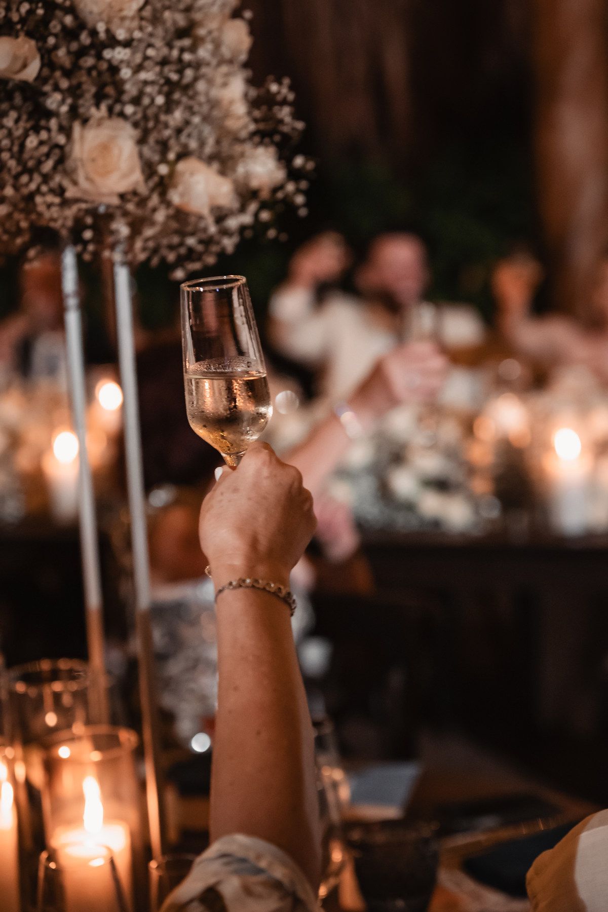 Guest raising champagne toast at candlelit wedding reception with floral centerpieces and warm glow.