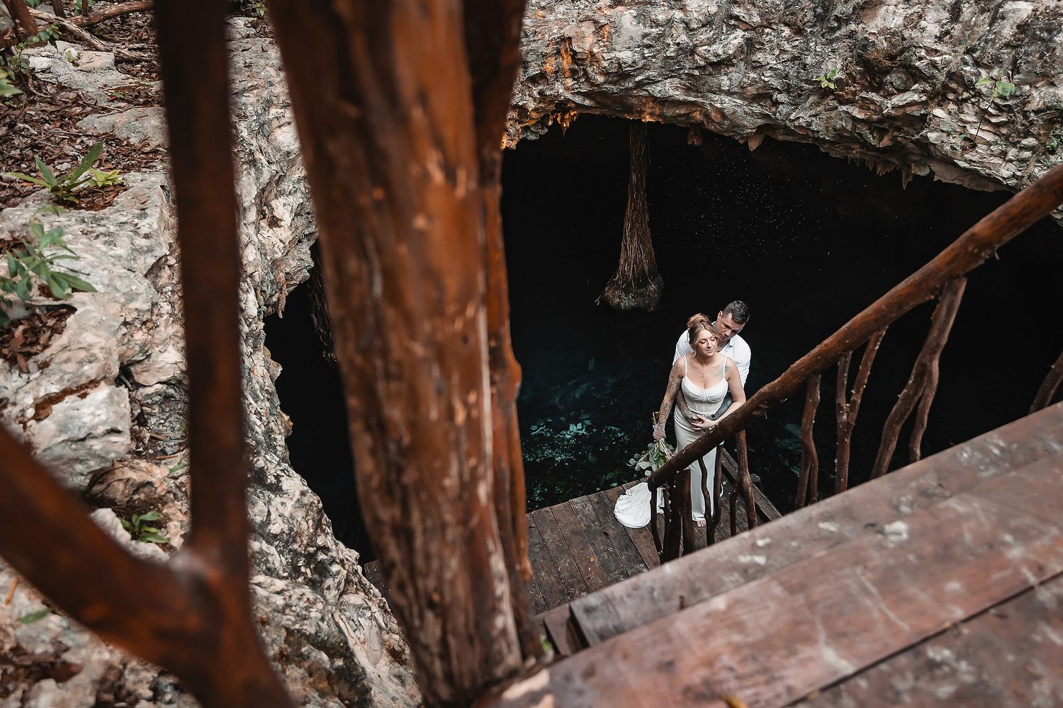 Couple sharing a romantic kiss on rustic stairs descending into a stunning cenote cave in Mexico.