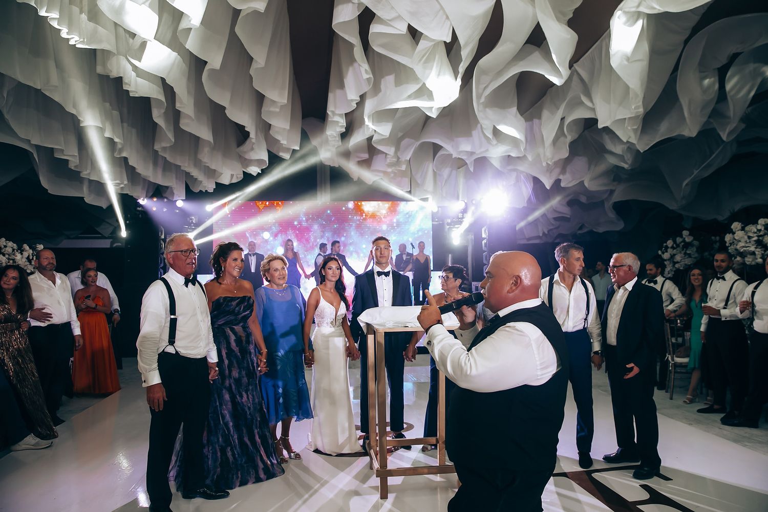 Wedding guests dance under elegant white paper lanterns and colorful lighting at an indoor reception celebration.
