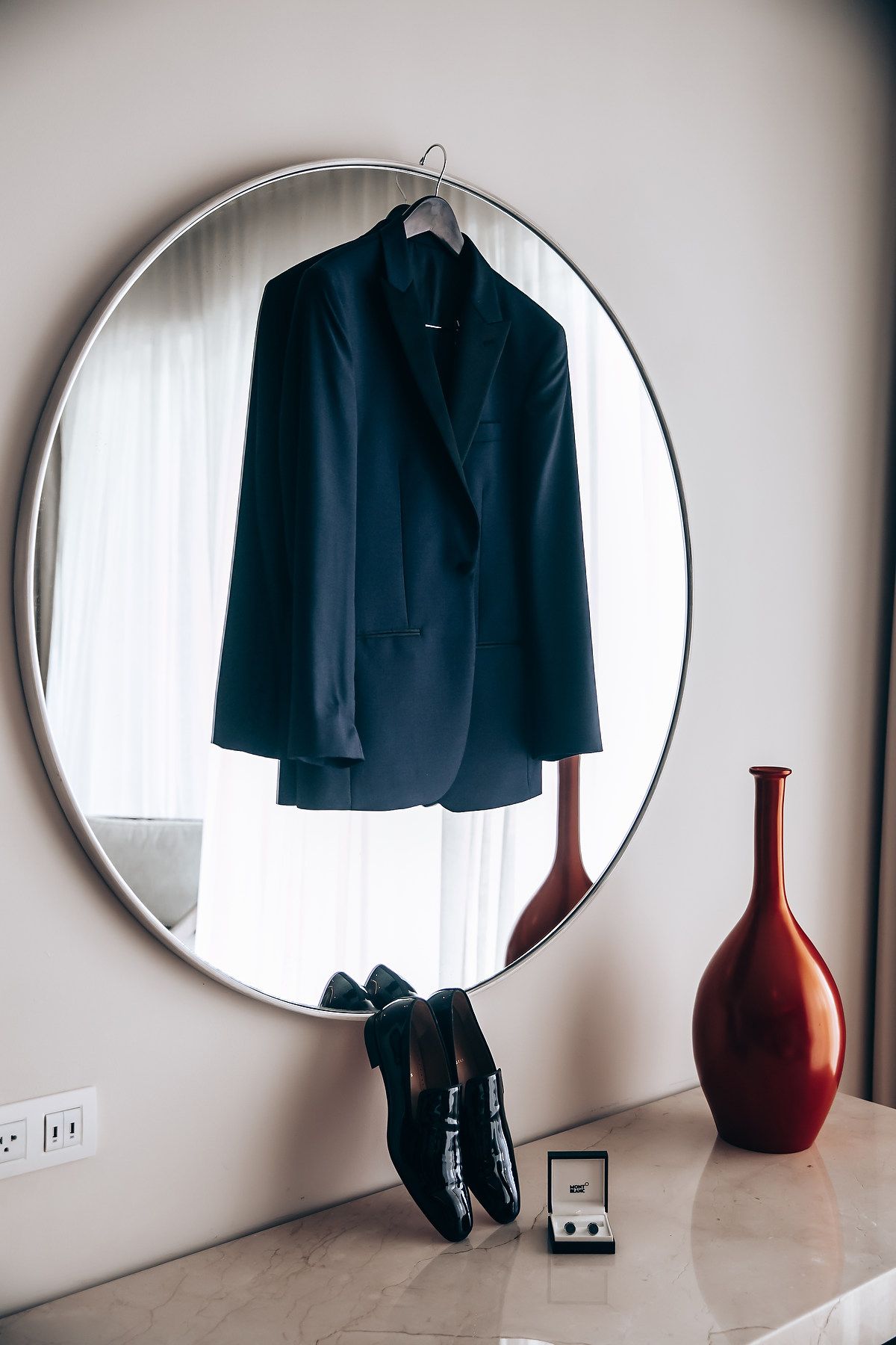 A black suit jacket and dress shoes displayed beside an oval mirror and red vase.