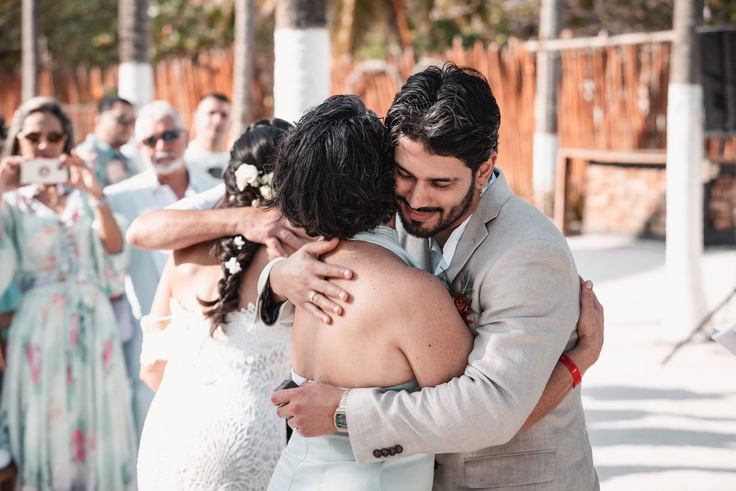 Bride and groom sharing emotional group hug with guest at tropical beach wedding celebration.