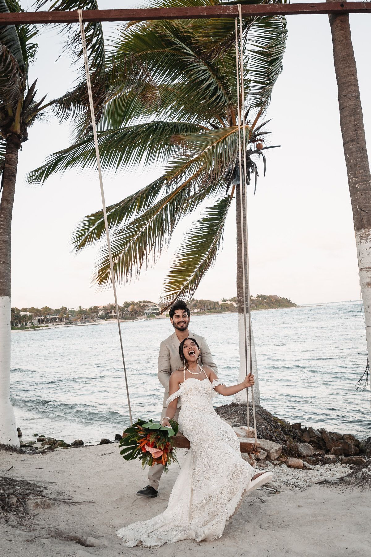Joyful bride and groom on a swing by the ocean under tropical palm trees.