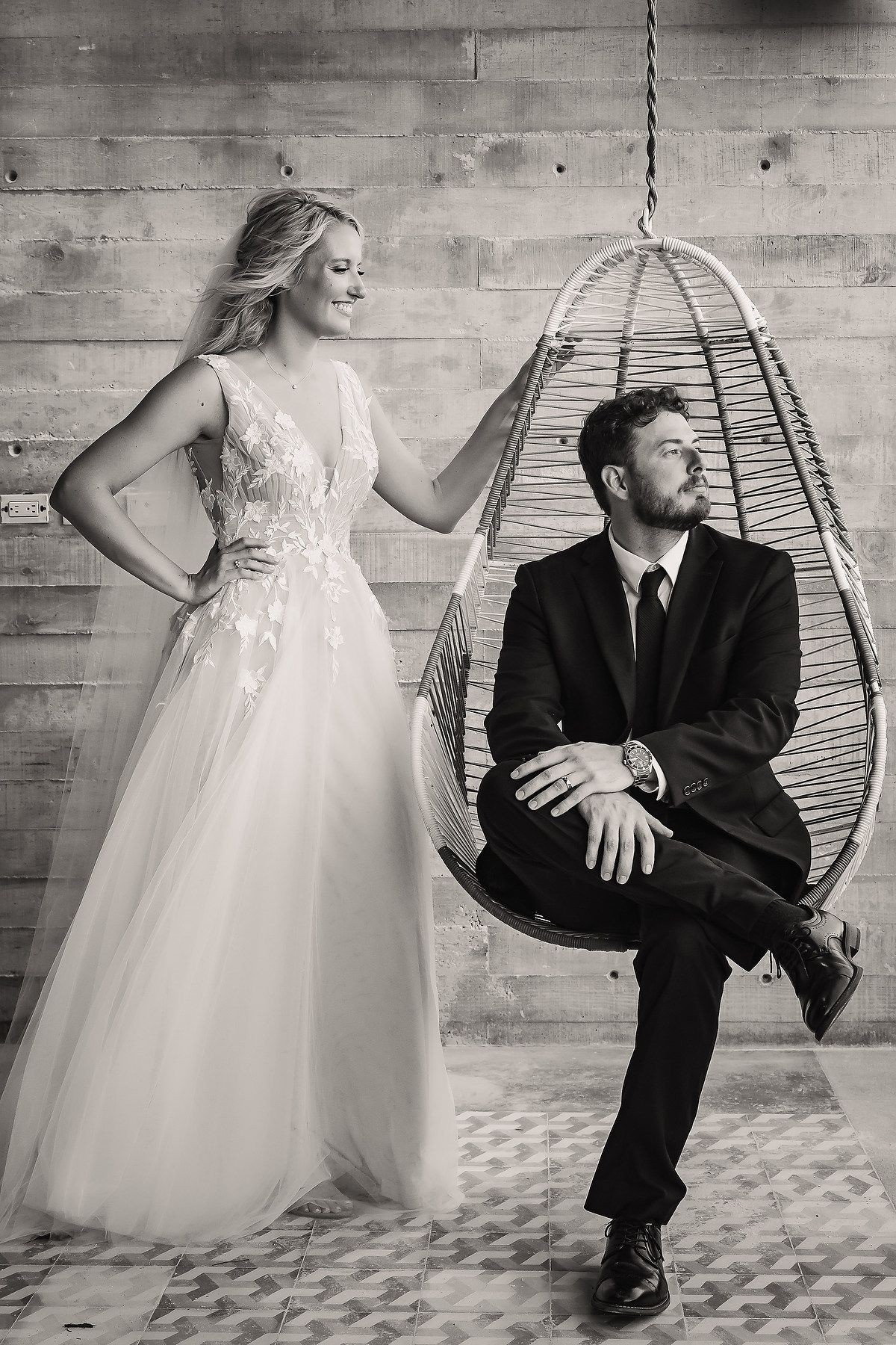 Black and white wedding photos show a couple posing with a hanging chair against a concrete wall.