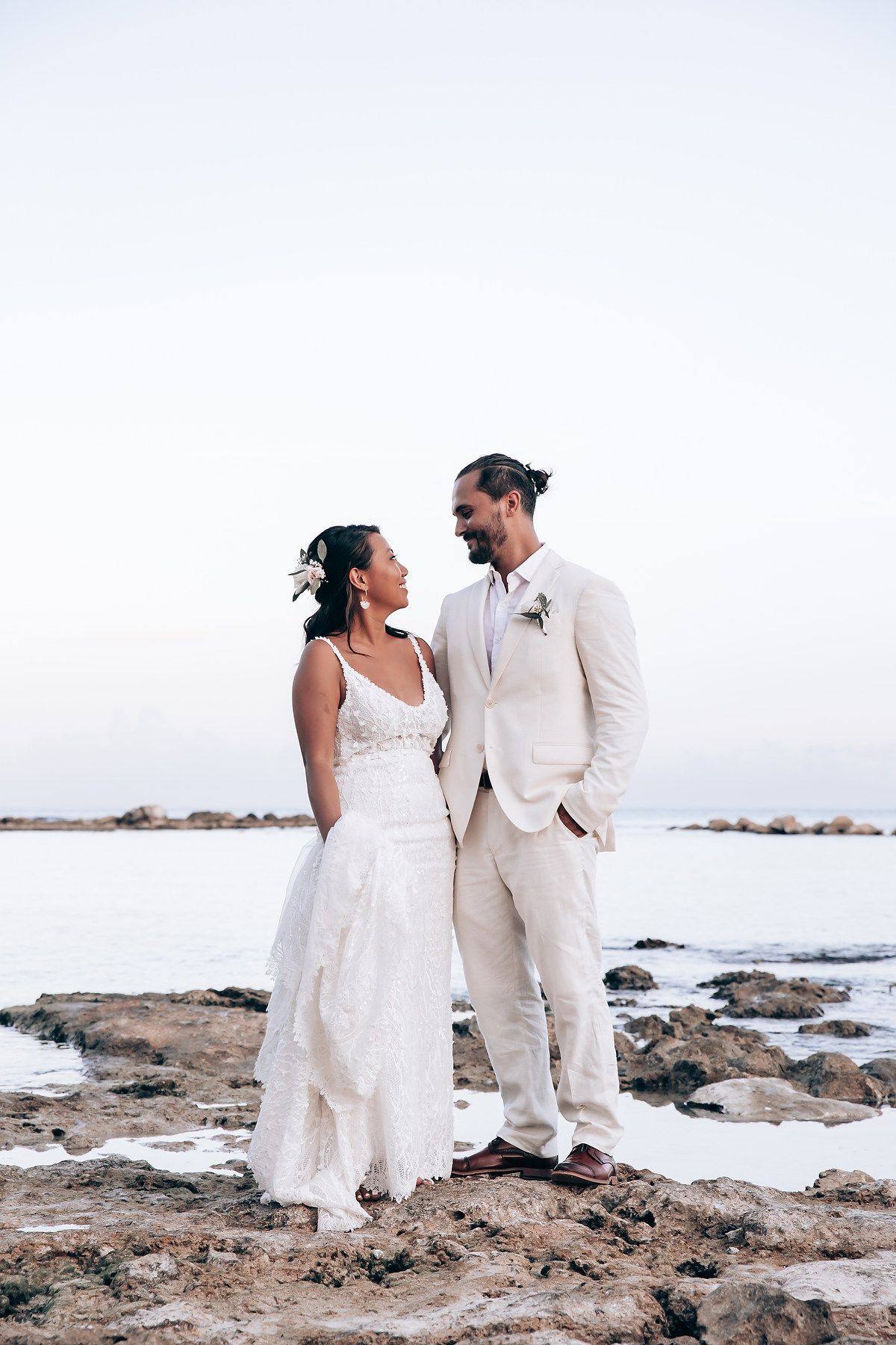 Couple in white attire walking along rocky coastal shoreline during sunset wedding photoshoot.