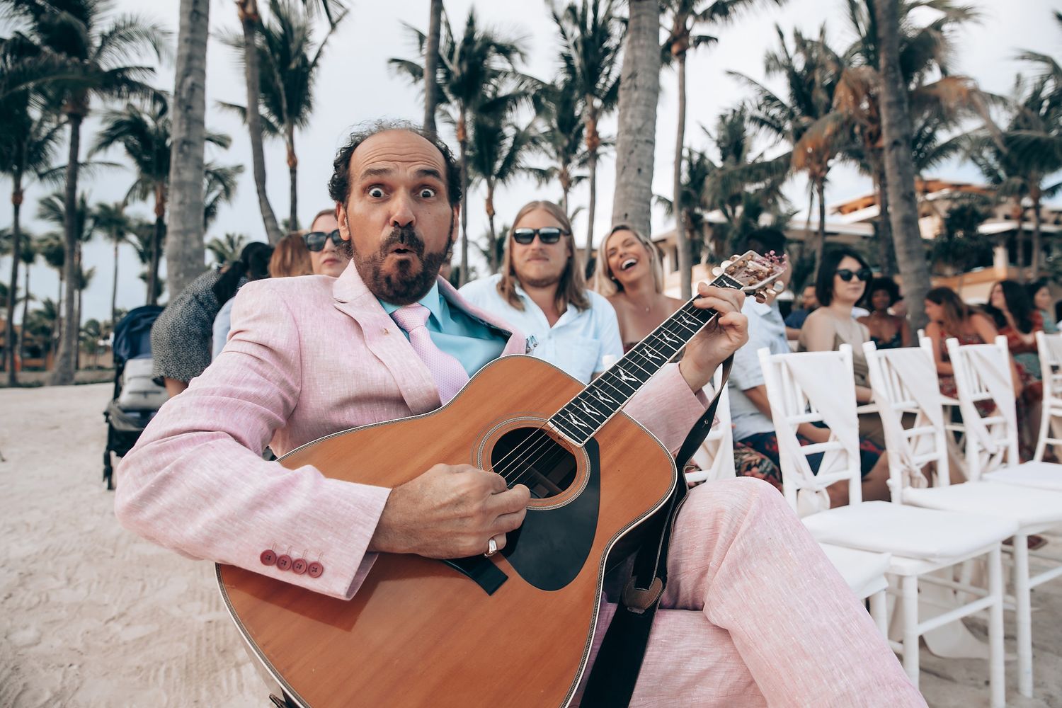 Musicians performing with guitars at a beach wedding ceremony with palm trees in background.
