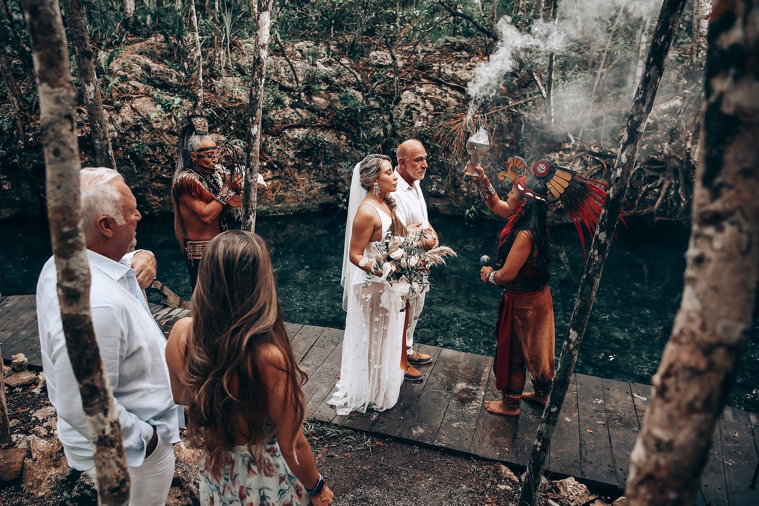 Wedding party gathers in a wooded outdoor setting for a ceremony with guests watching in casual attire.