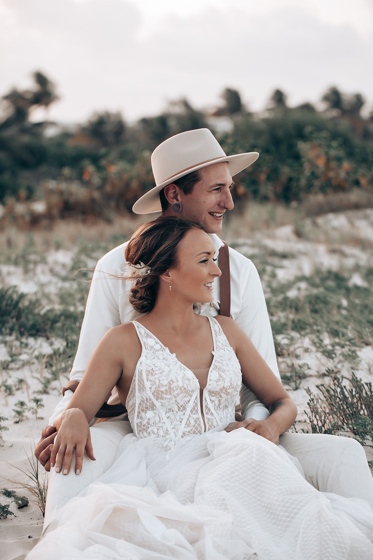 Couple laughing together while sitting in white attire on a sandy beach with coastal vegetation.