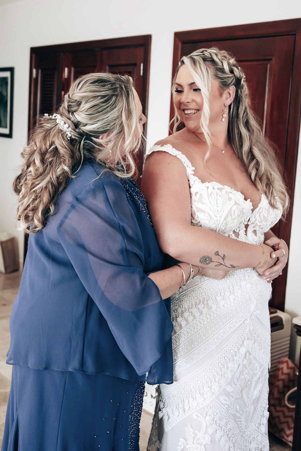 A bride smiles as a woman in blue helps fasten her lace wedding gown indoors.