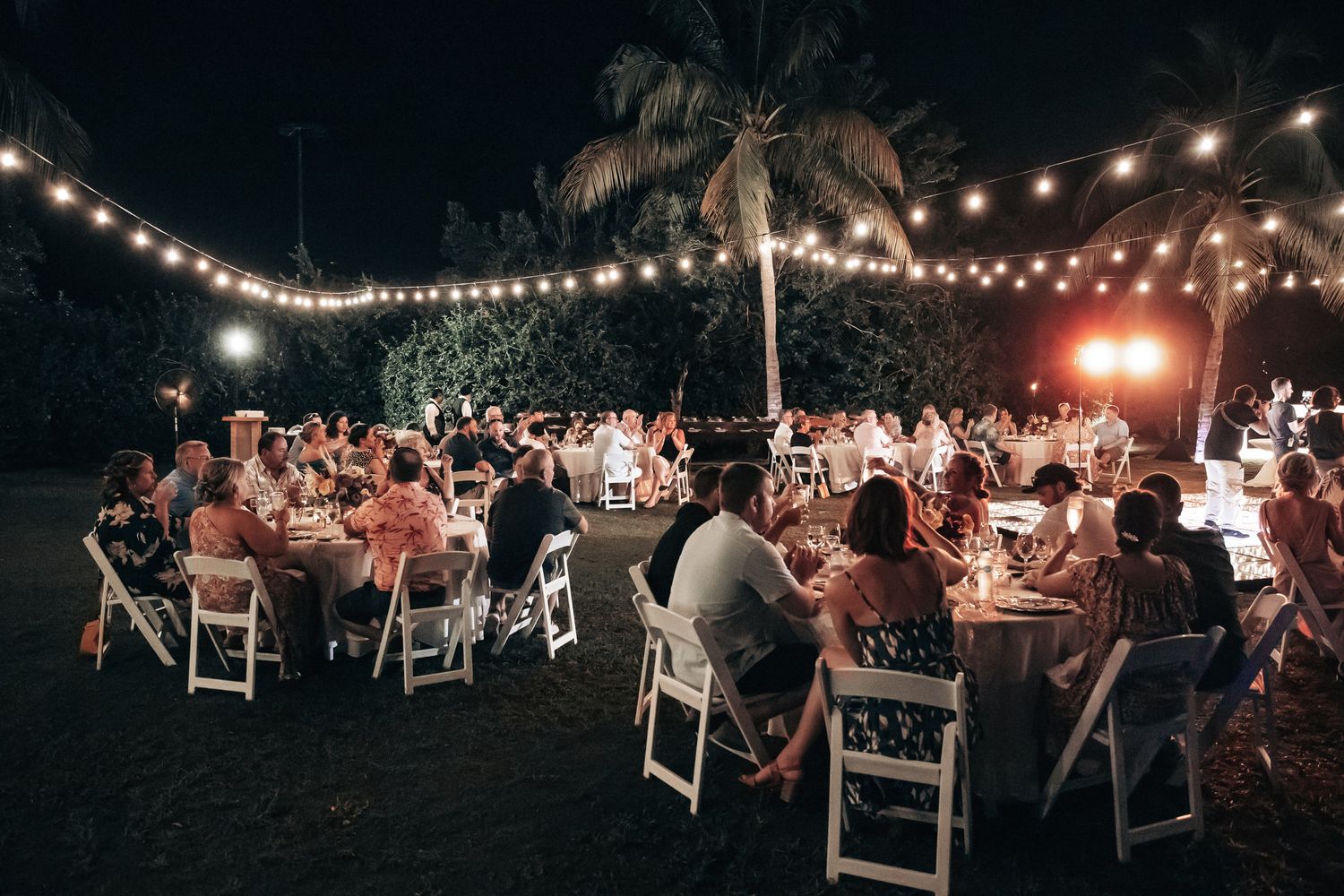 Guests dining under string lights at an elegant outdoor tropical wedding reception at night.