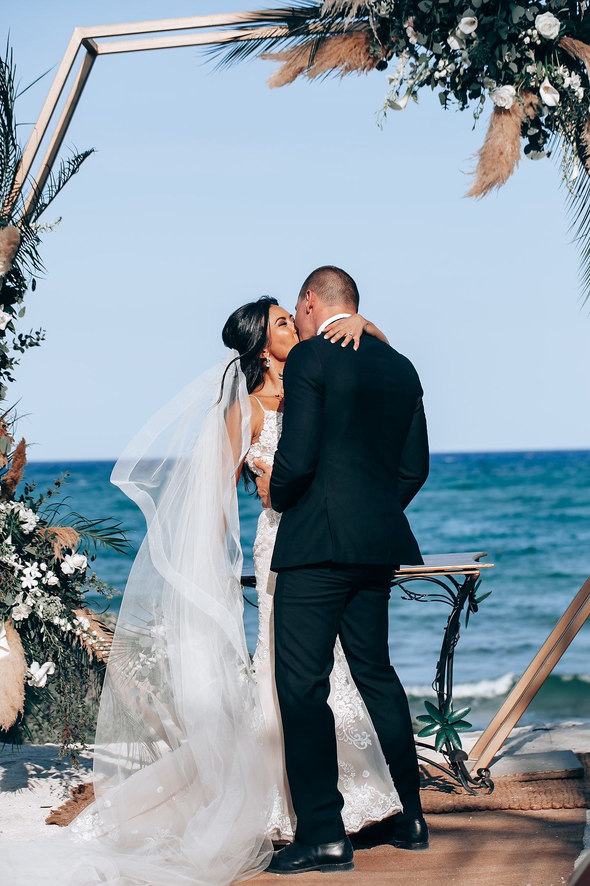 Intimate beach wedding ceremony with wooden arch and white floral decorations against turquoise waters.