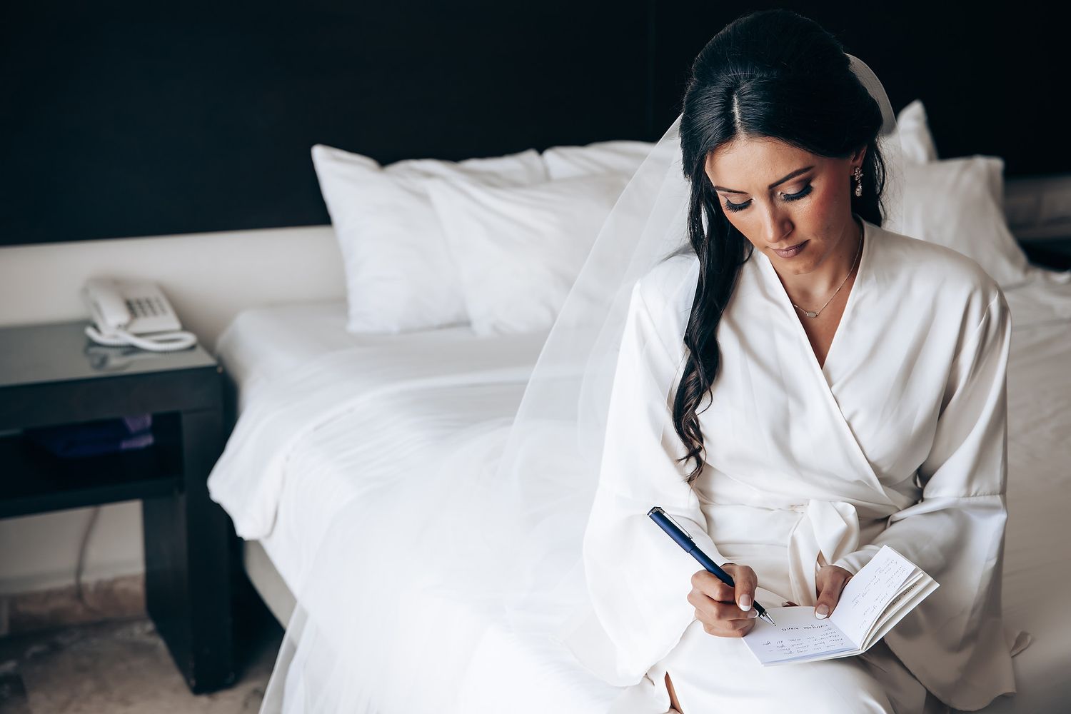 Someone in a white robe sits on a bed writing in the morning light.