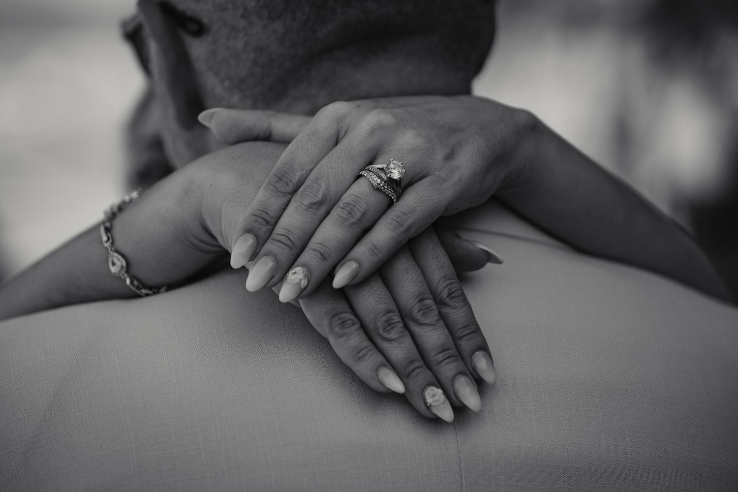 Close-up black and white photo of bride's hands with diamond ring and bracelet resting on groom's back.