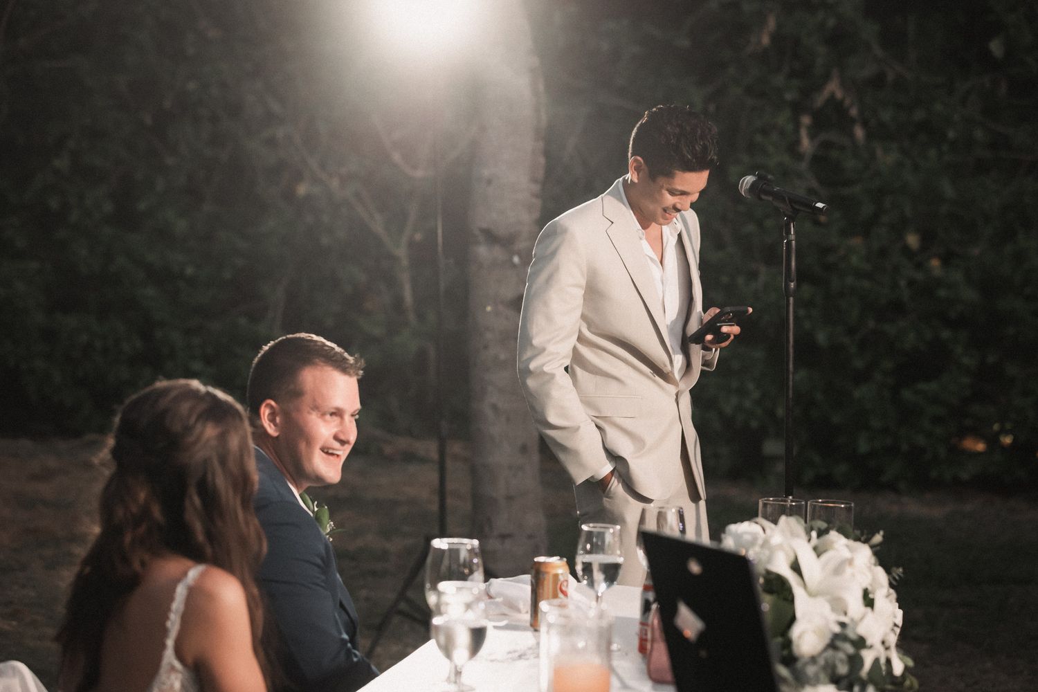 Guest delivers heartfelt speech to smiling bride and groom at outdoor evening wedding reception dinner.