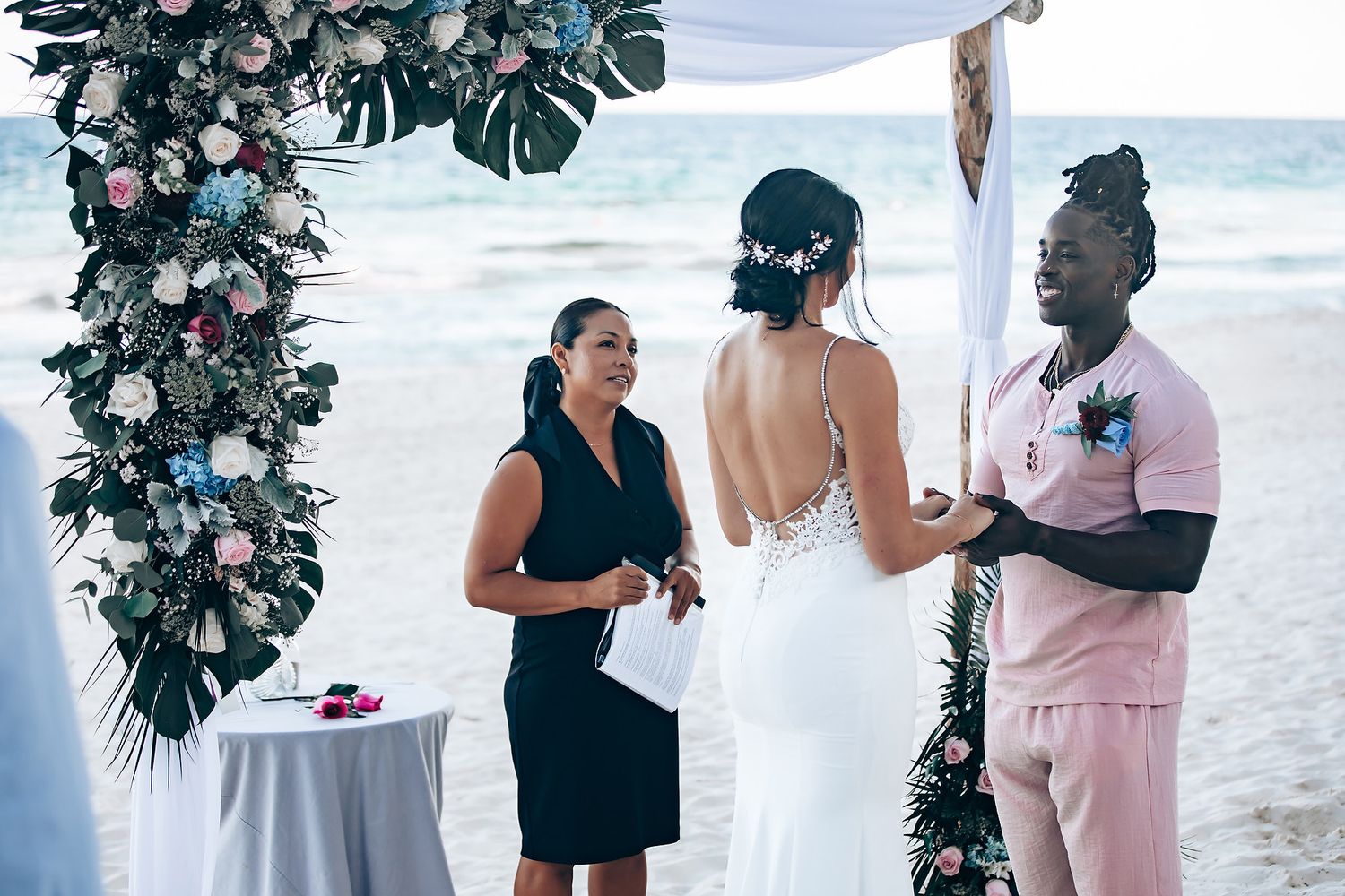 A couple embraces at a tropical wedding ceremony with a floral arch in the background.
