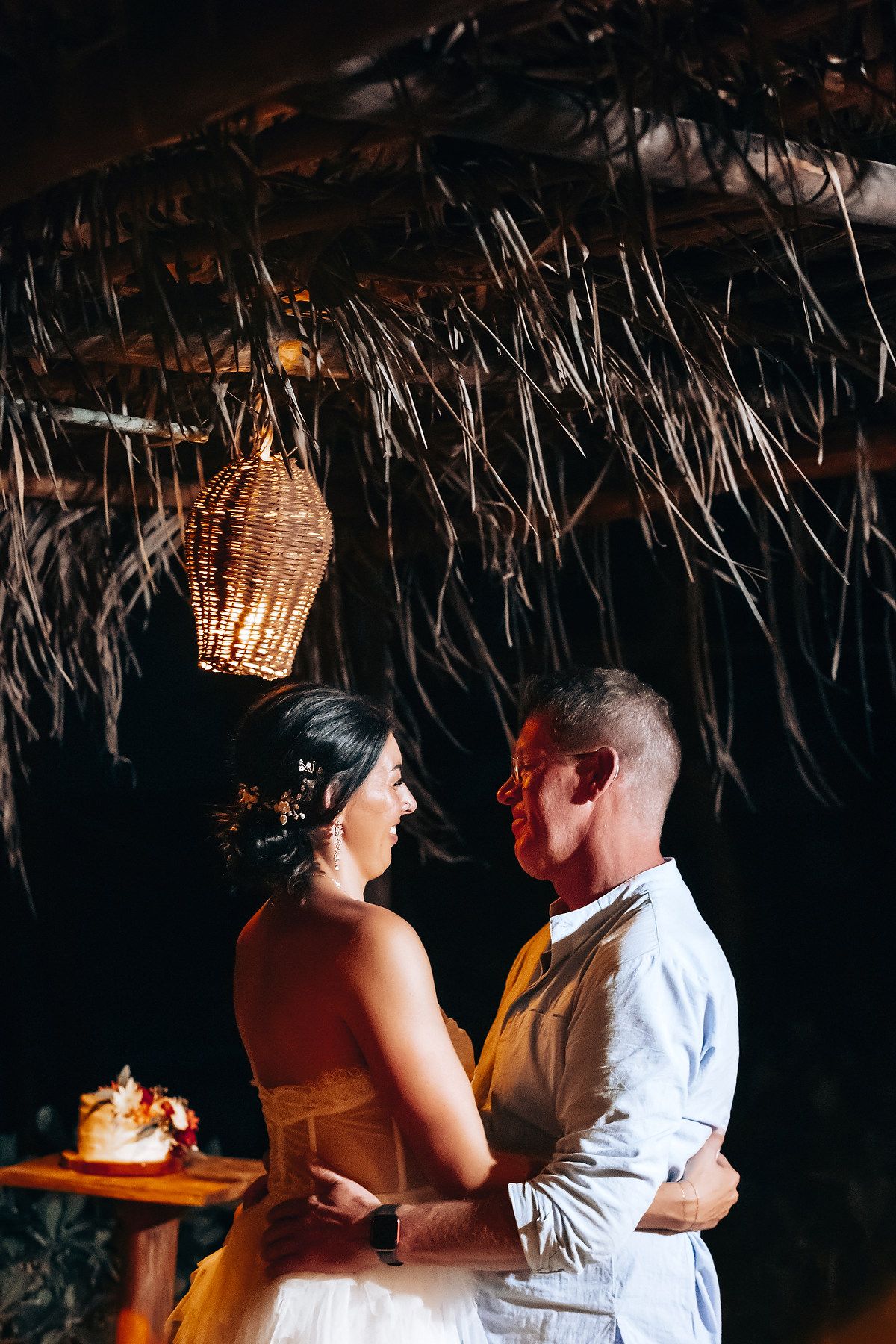 A couple embraces at their tropical wedding reception, surrounded by lush greenery and hanging lanterns.