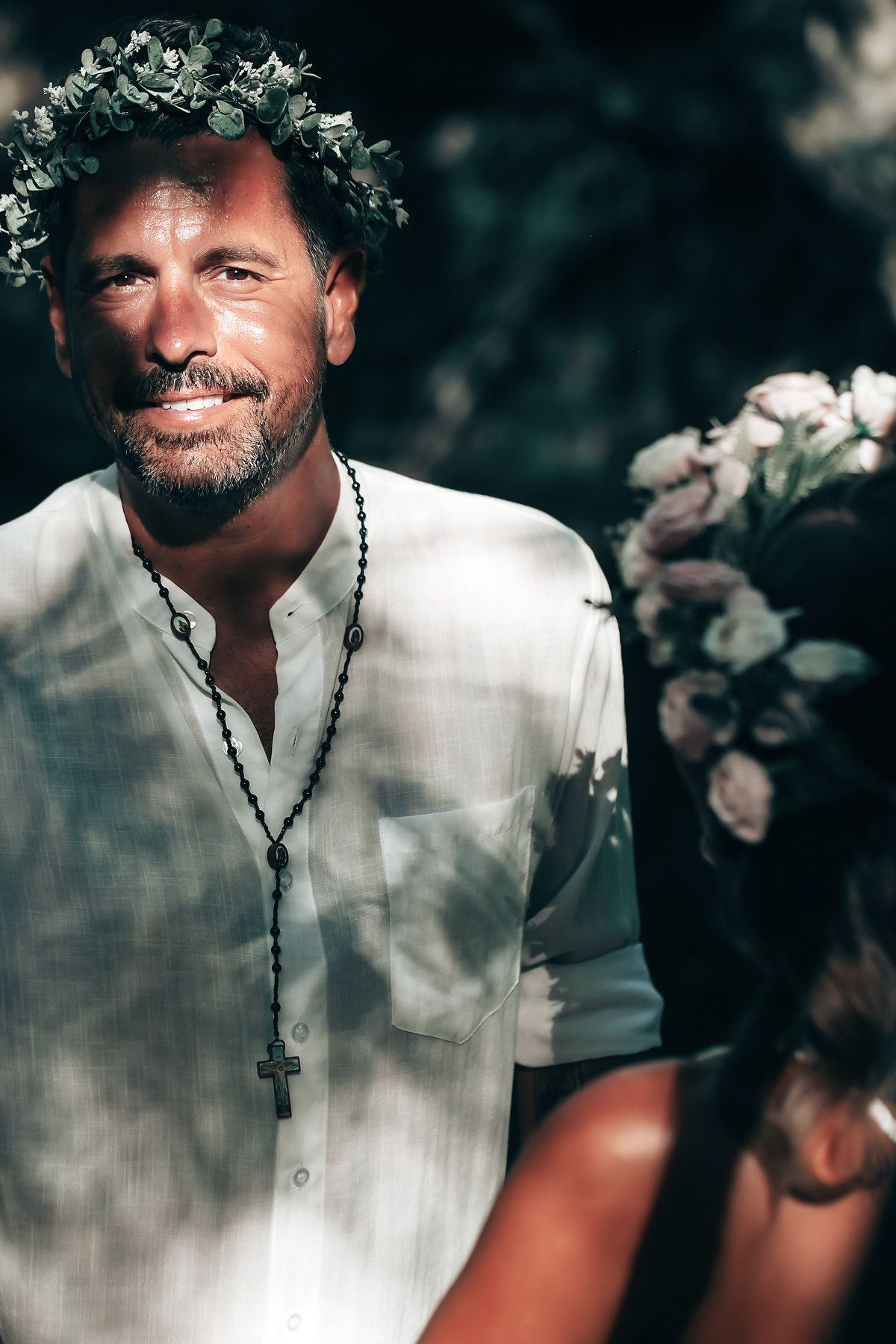 A man with a beard wearing a white shirt and a floral wreath on his head stands in a garden.