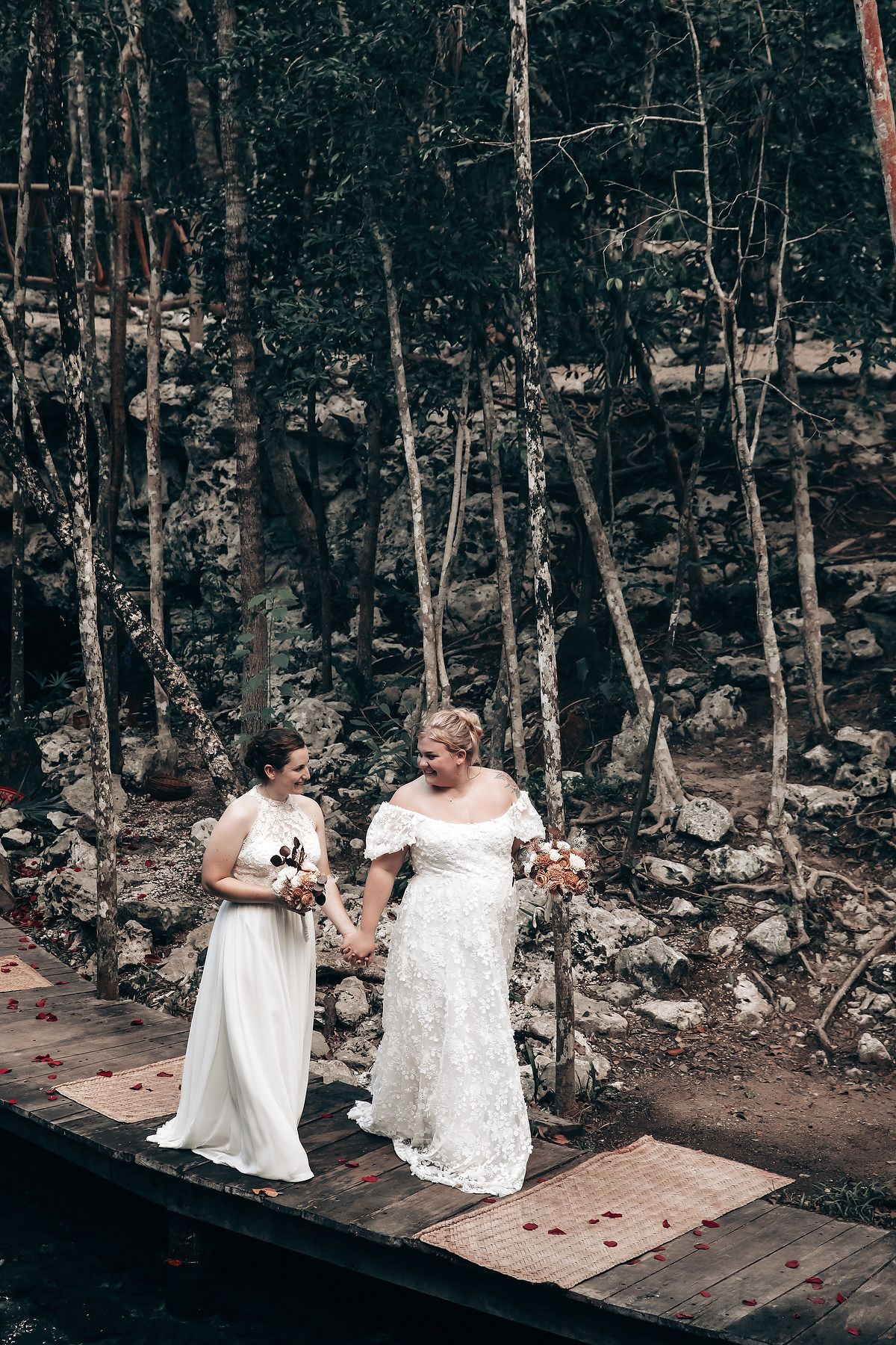 Two brides in white wedding dresses standing on a wooden walkway in a tropical forest.