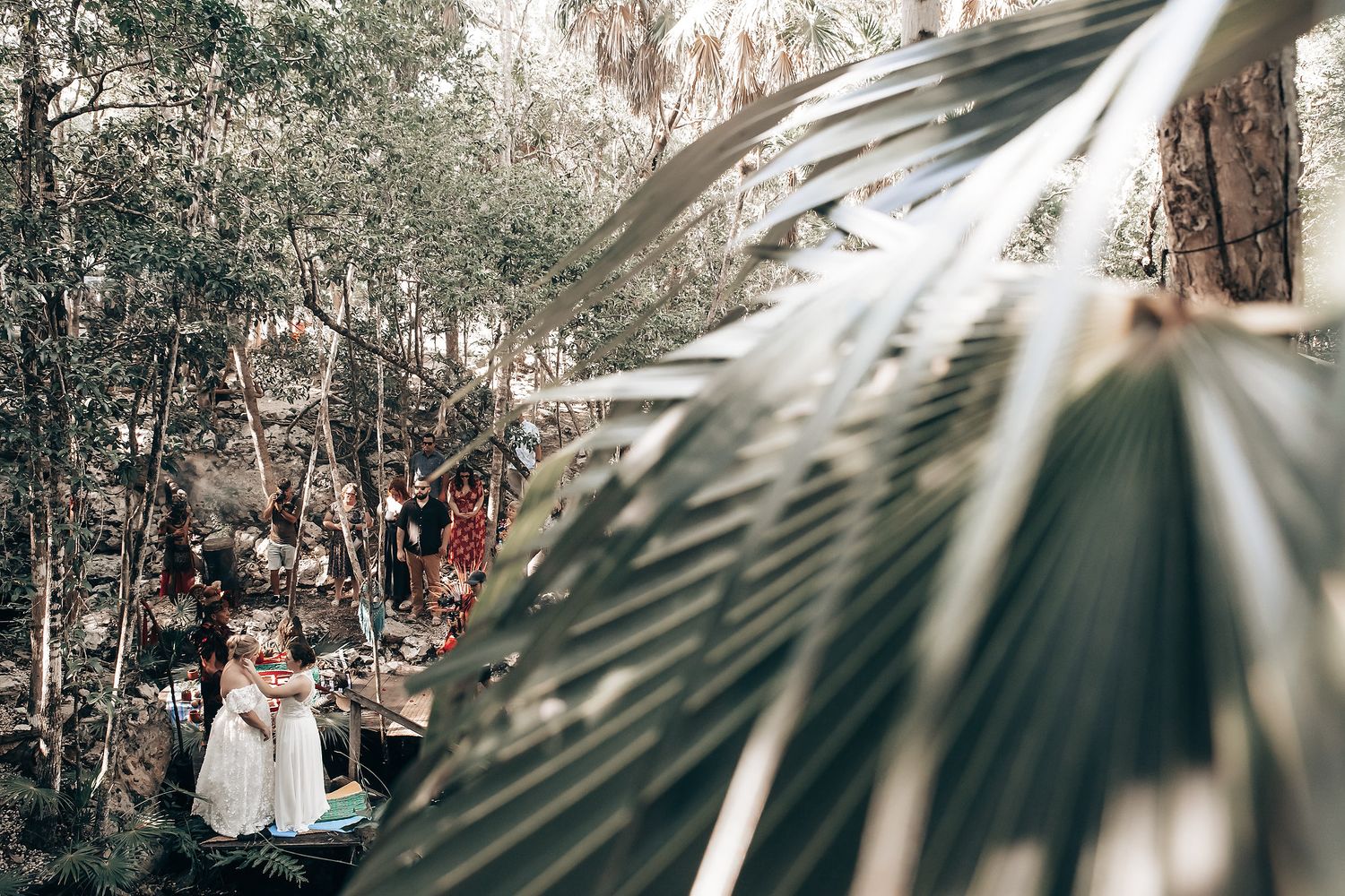 A person wearing a white dress stands in a tropical forest setting with palm fronds in the foreground.