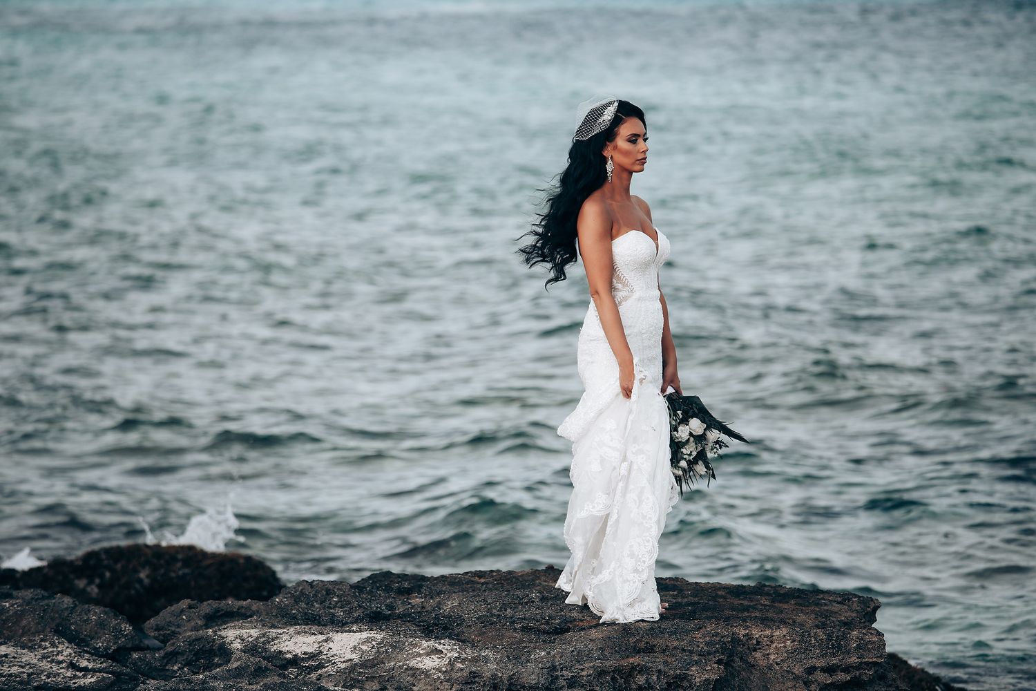 Bride in white wedding dress poses on rocky coastline near ocean waves.