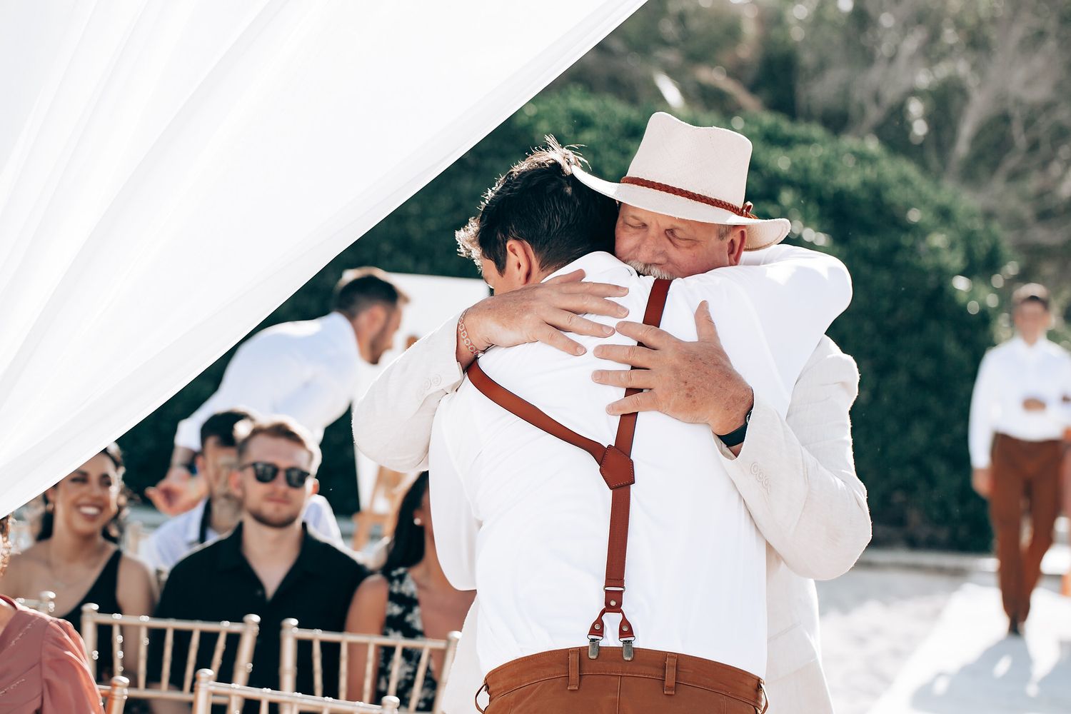 Wedding guests in white attire and suspenders gather outdoors under white canopy decorations.