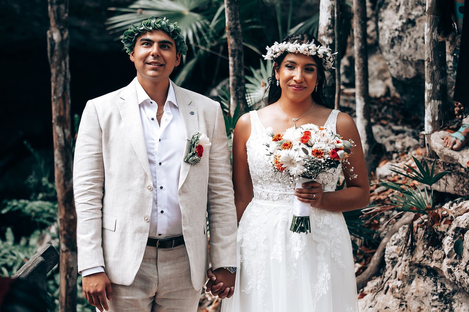 A wedding couple poses together in a wooded outdoor setting wearing white attire and floral accessories.