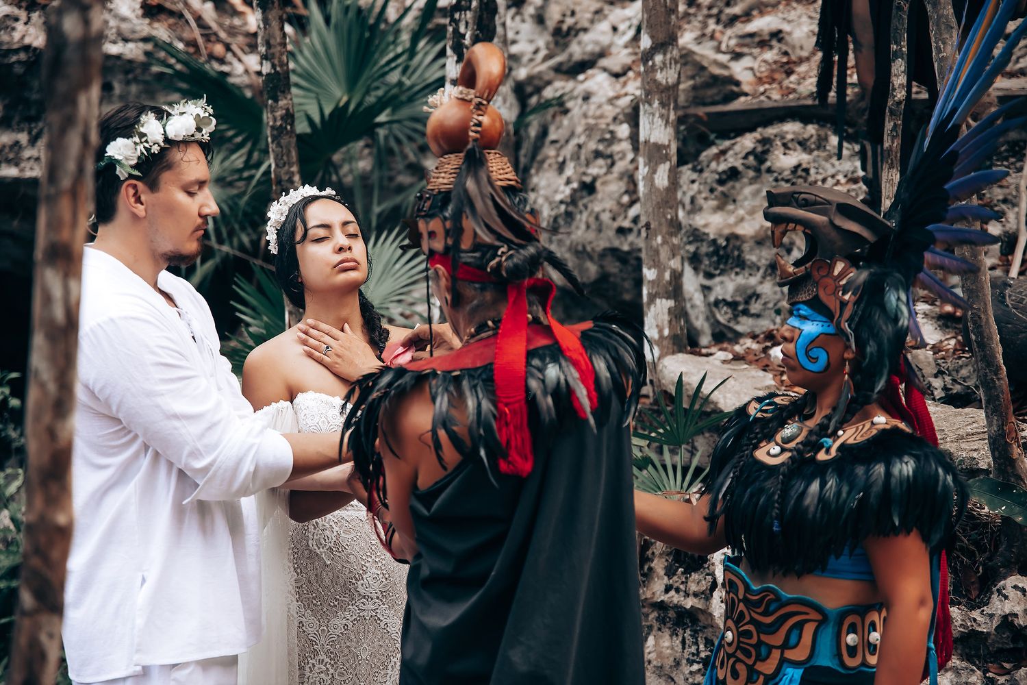 Wedding guests gather outdoors near a stone wall with tropical plants during a celebration ceremony.