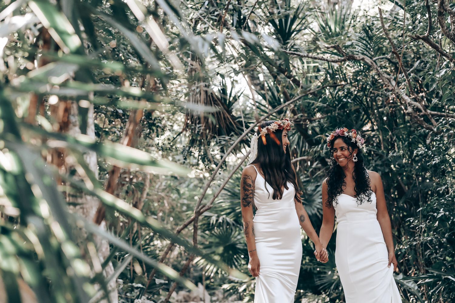 Two people in white dresses stand together in a tropical garden setting with palm trees.