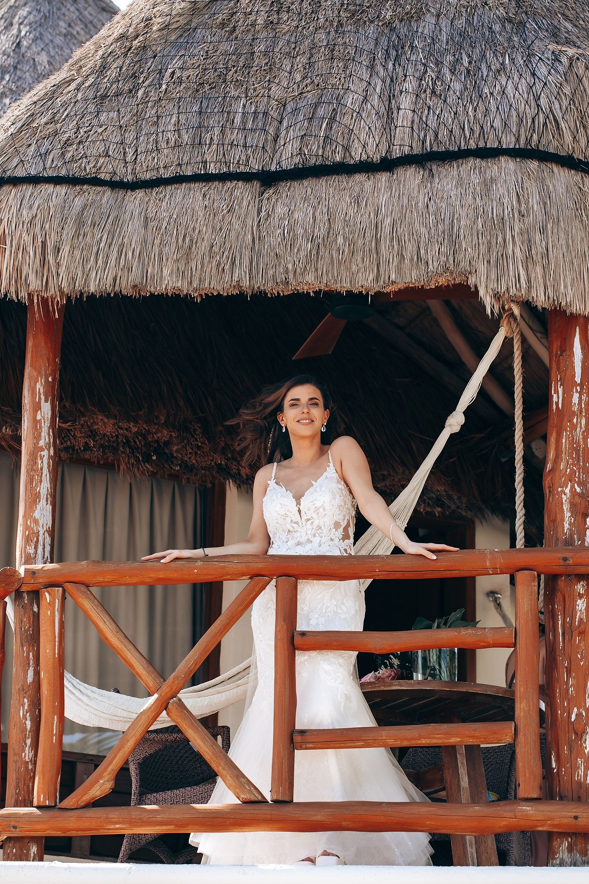 A white wedding dress is displayed in a tropical thatched-roof cabana overlooking the beach.