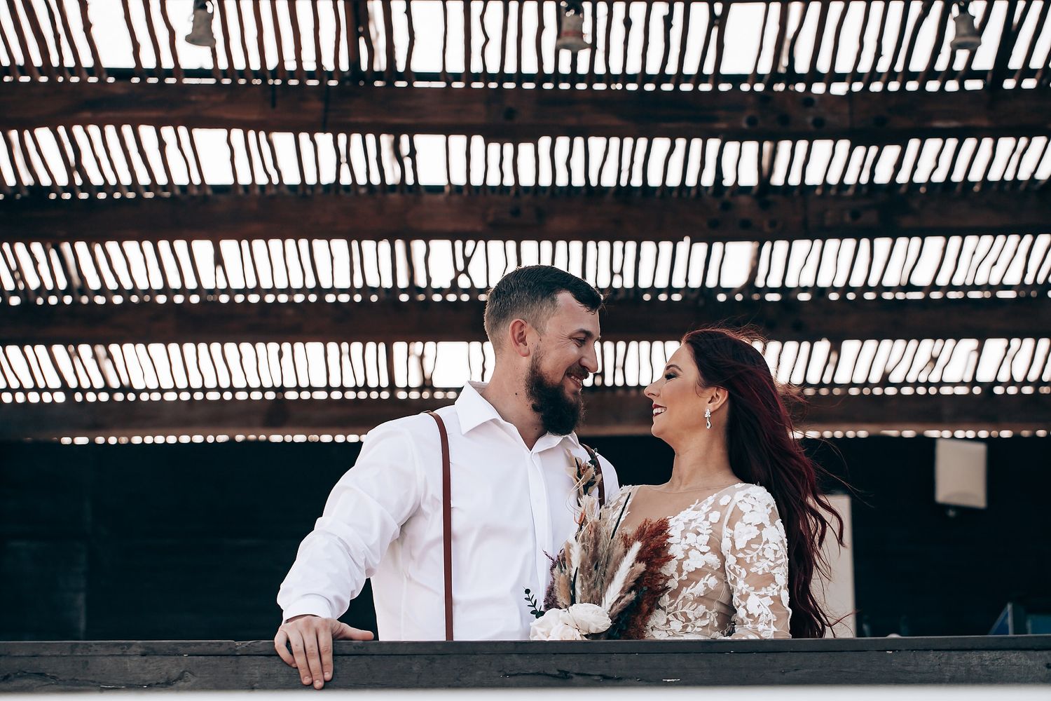 Wedding couple embracing under a rustic wooden pergola with slatted roof creating dramatic shadows.