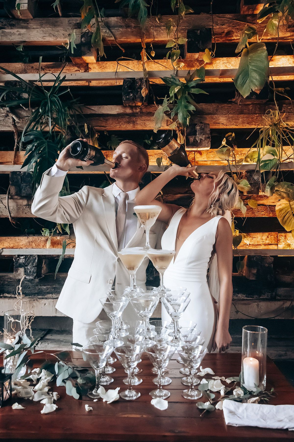 White-dressed figures posing with champagne glasses against rustic wooden backdrop with floral decorations.